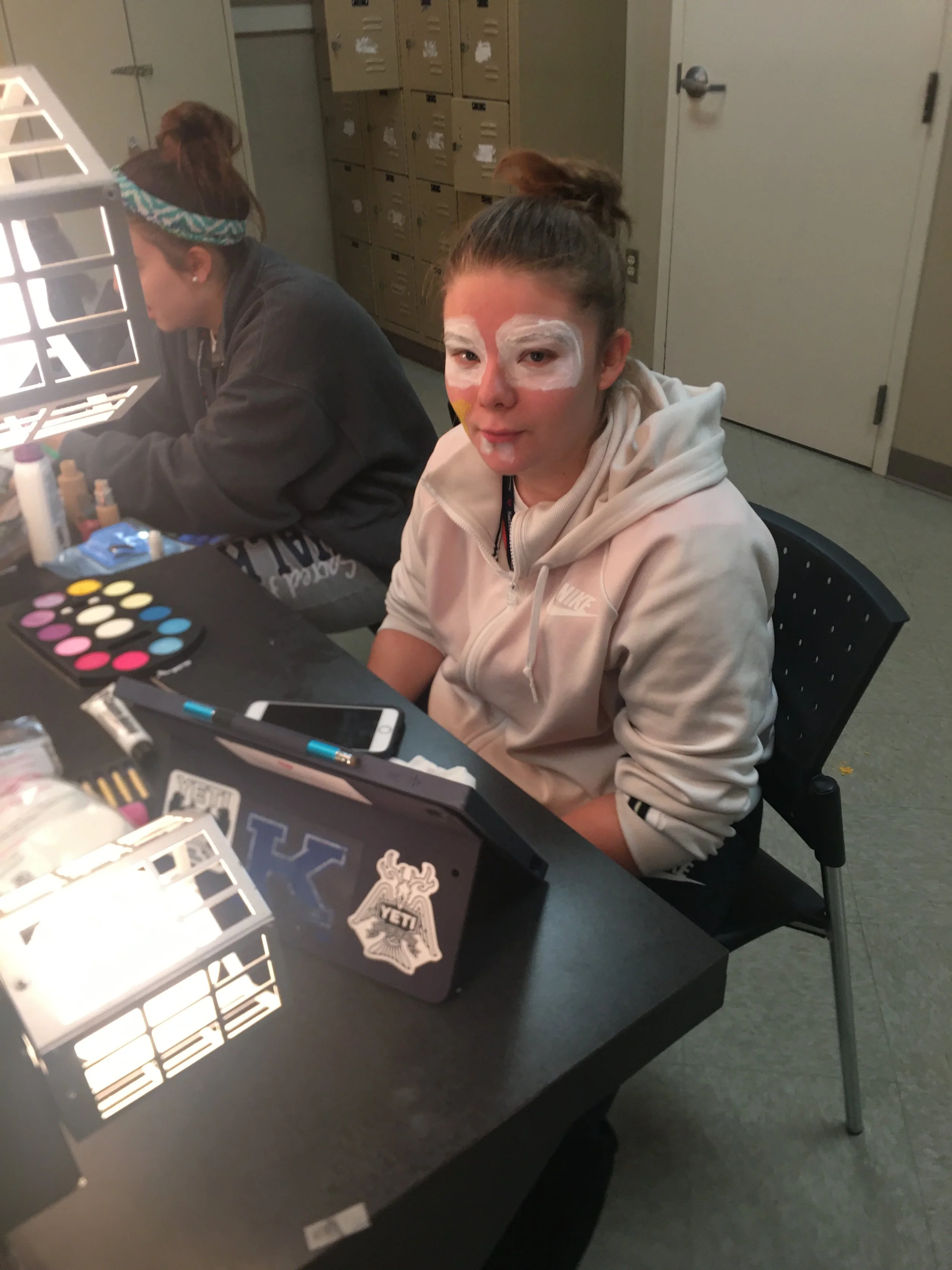 A young woman with face paint sitting at a table with makeup supplies, a mirror, and a smartphone, in a room with lockers in the background.