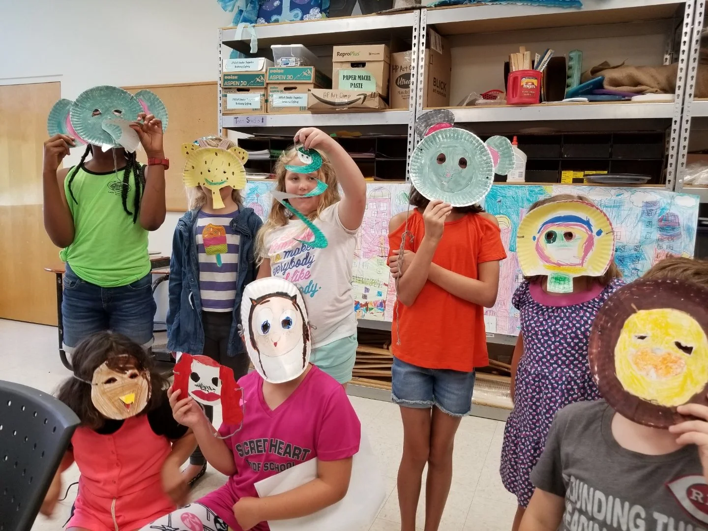 Children wearing paper plate masks shaped like various faces, in a classroom setting with shelves and artwork in the background.
