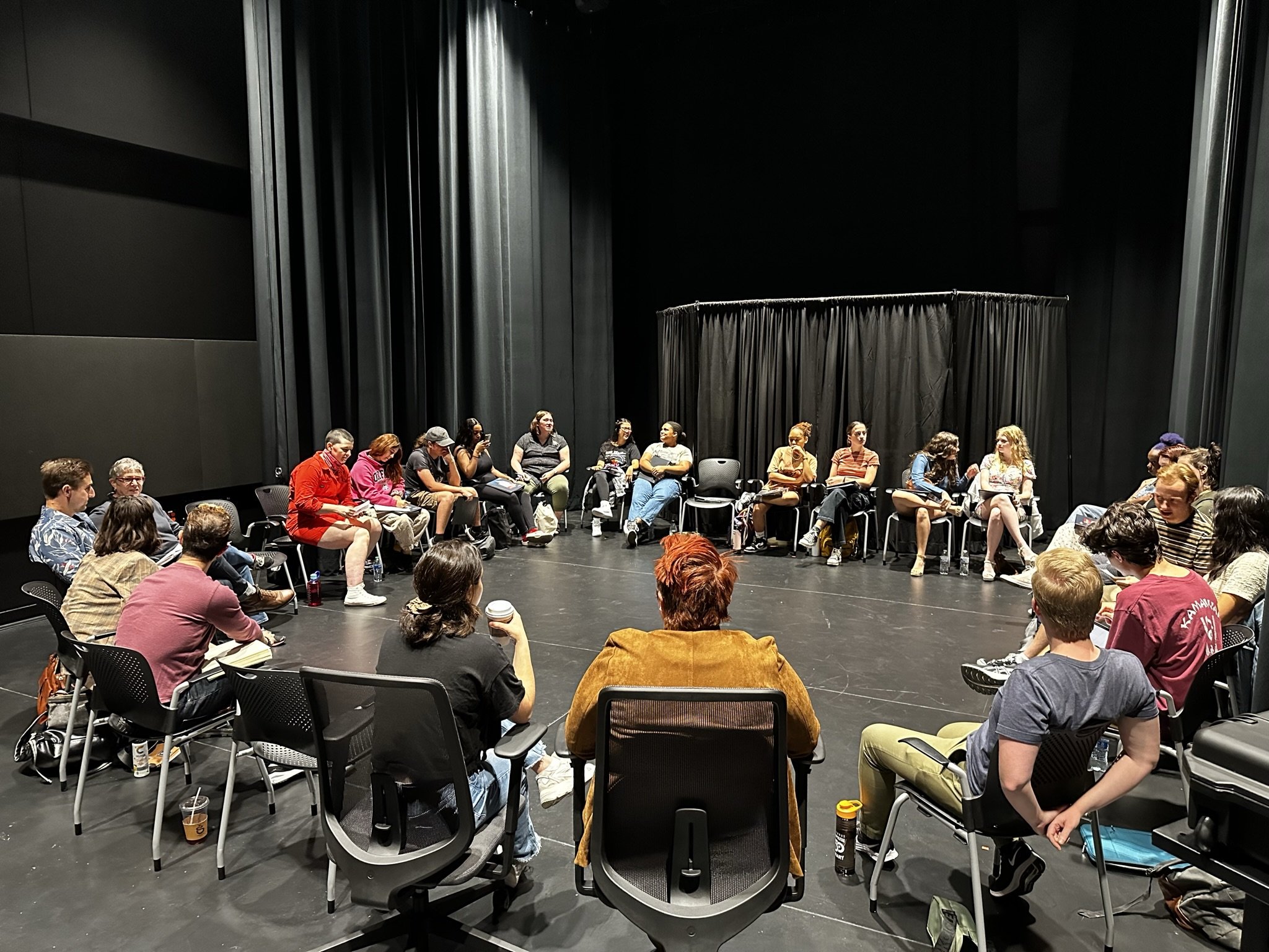 Group of people sitting in a circle on a black stage with black curtains, participating in a discussion or workshop.