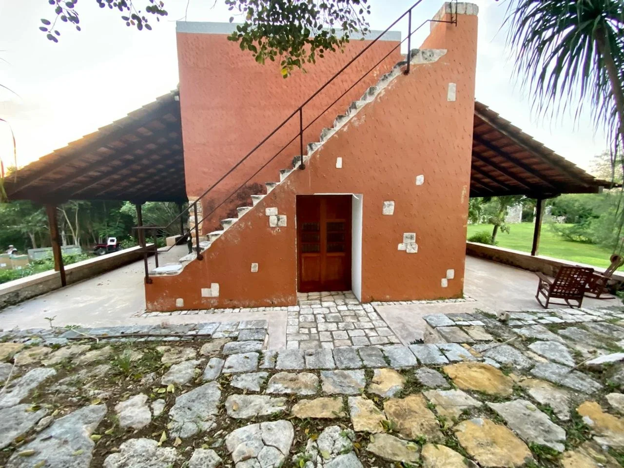 A rustic outdoor structure with a terracotta-colored wall and a staircase leading to a rooftop, surrounded by a stone pathway and outdoor seating, with green trees and grassy landscape in the background.