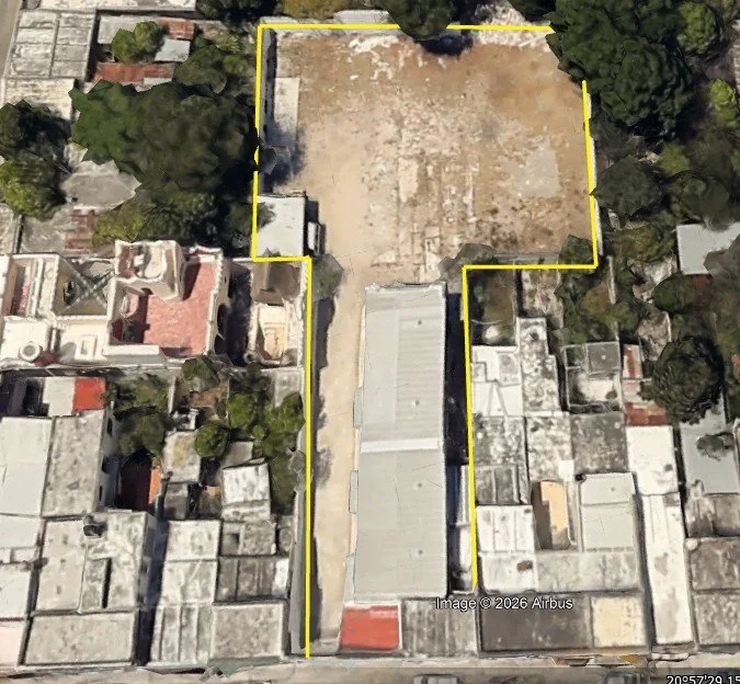 An aerial view of a property outlined in yellow, showing surrounding houses and trees, with a large open area of bare ground.
