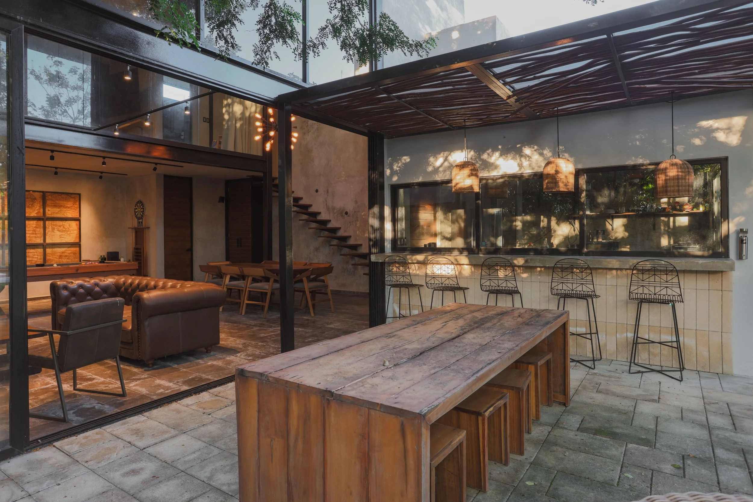 Outdoor patio area with a wooden table and benches, barstools at a bar counter, hanging pendant lights, and a view into an indoor lounge with leather chairs, a dining table, and warm lighting.
