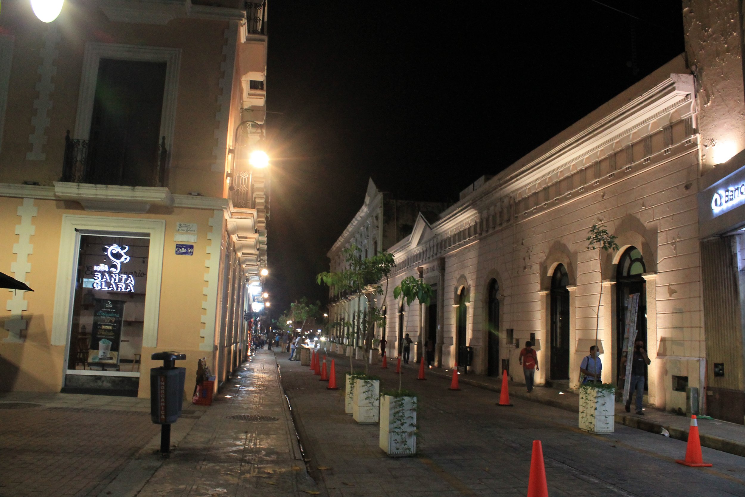 A nighttime street scene with historic buildings, streetlights, small trees in planters, and orange traffic cones along the sidewalk.