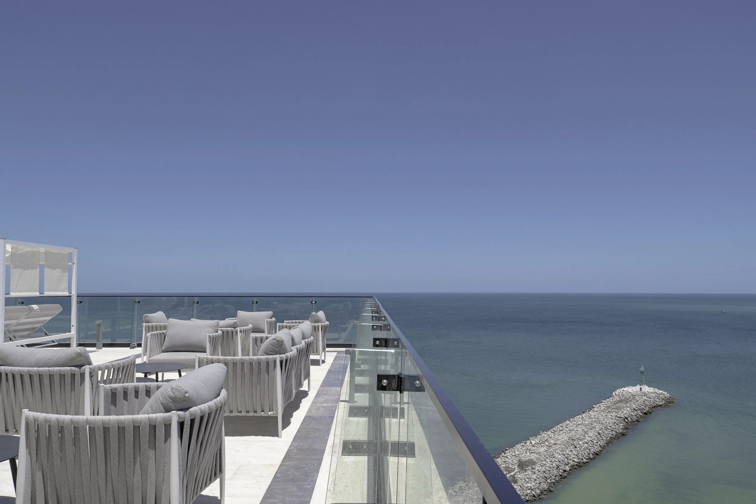 Rooftop terrace with outdoor seating and a glass railing overlooking the ocean, with a breakwater and lighthouse in the distance under a clear blue sky.