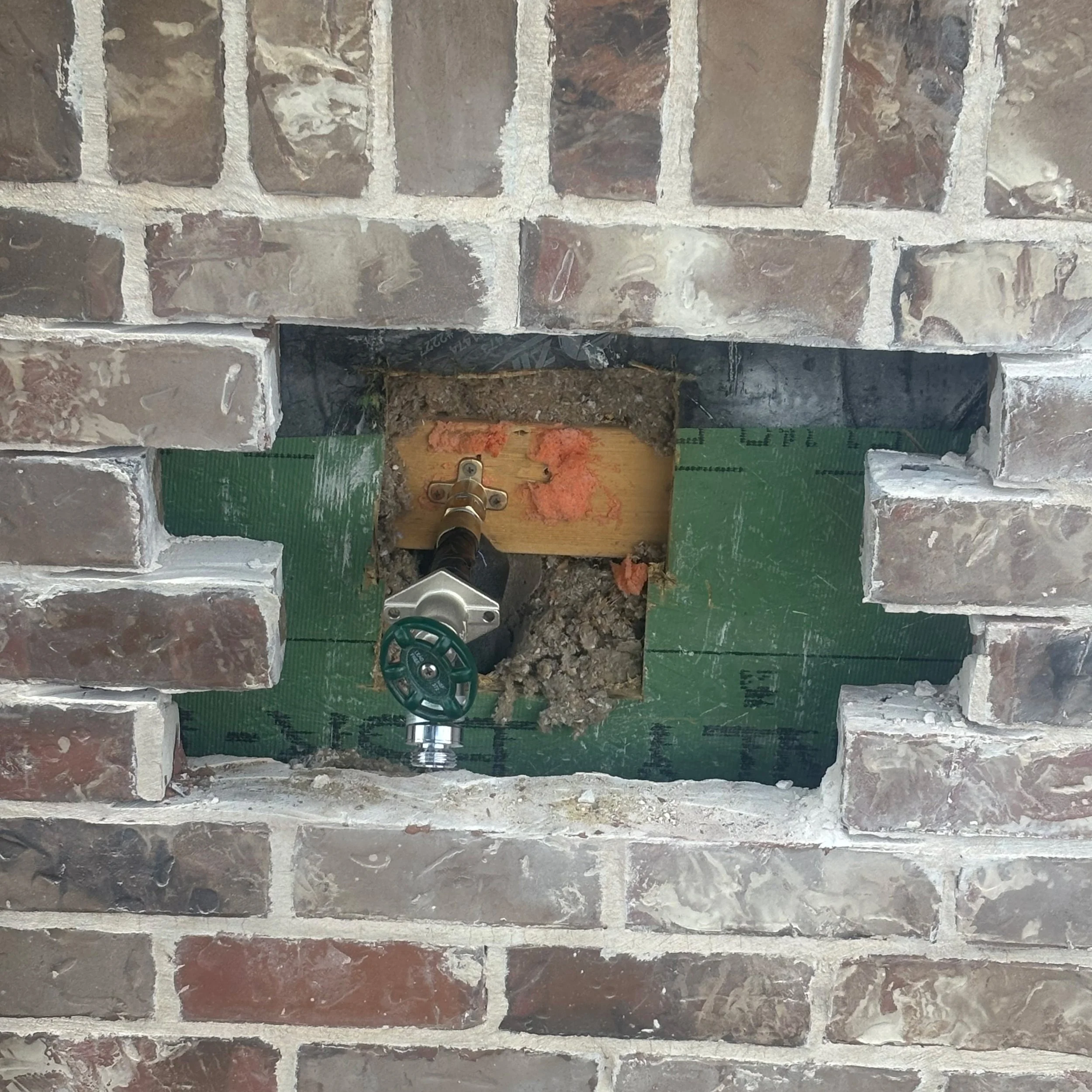 Exterior hose bib replacement through brick veneer, showing new valve, sheathing, and weather barrier before masonry repair