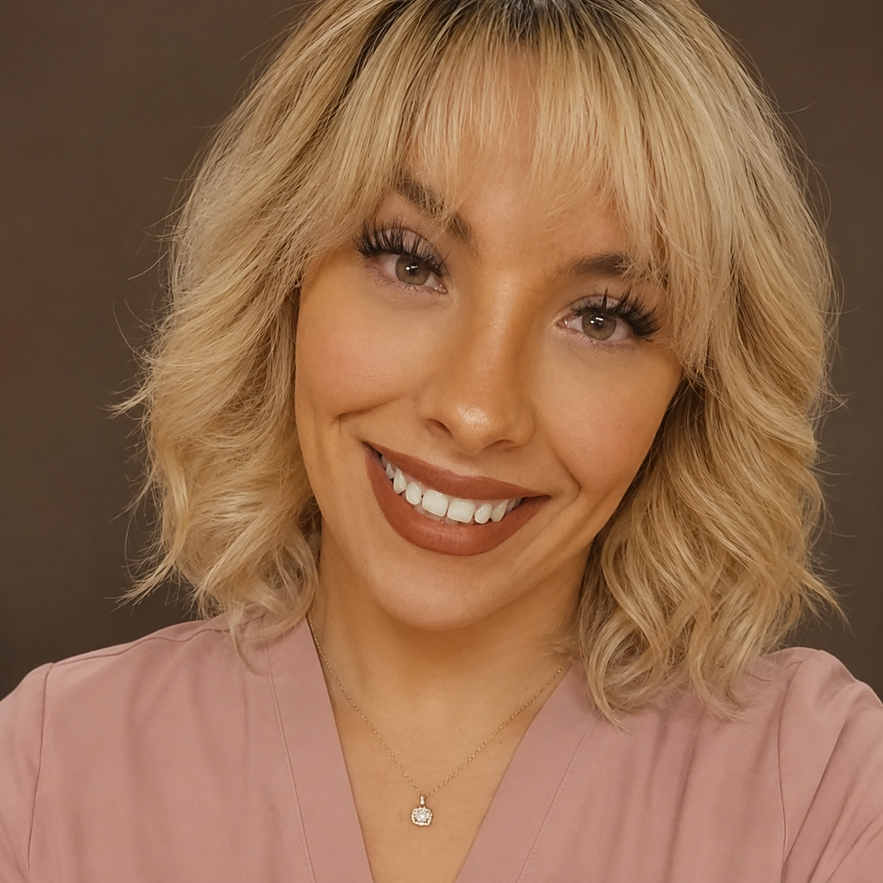 Portrait of a young woman with long brown hair, wearing a gray blazer and black top, smiling in front of a light gray background.