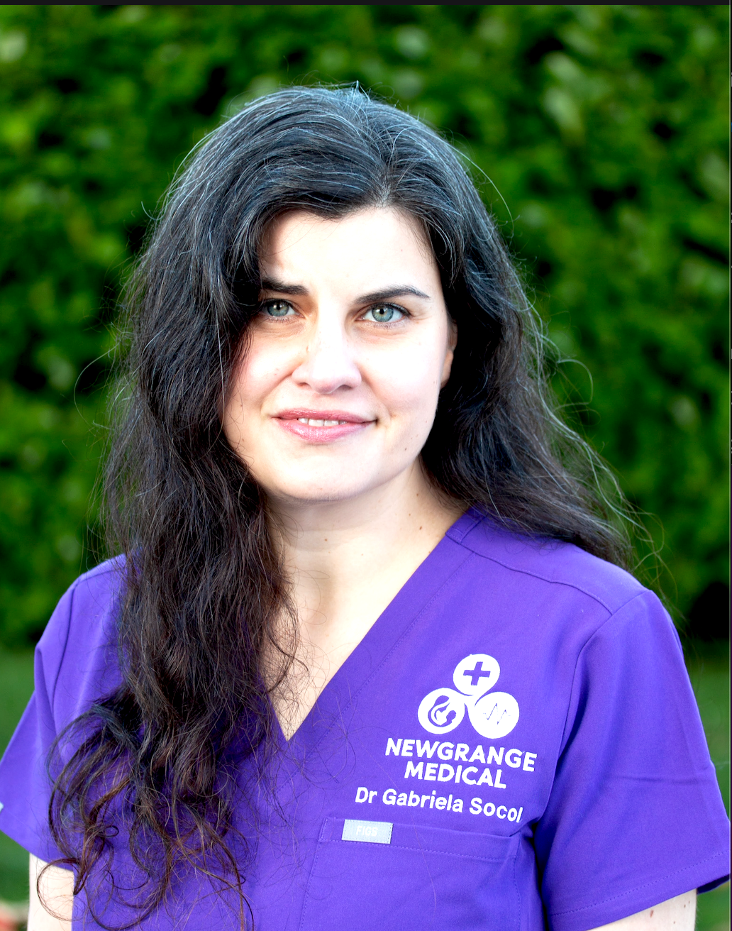 A woman with long dark hair, wearing a purple medical uniform with the text "Newgrange Medical, Dr. Gabriela Soco" and medical symbols, stands outdoors with a green leafy background.