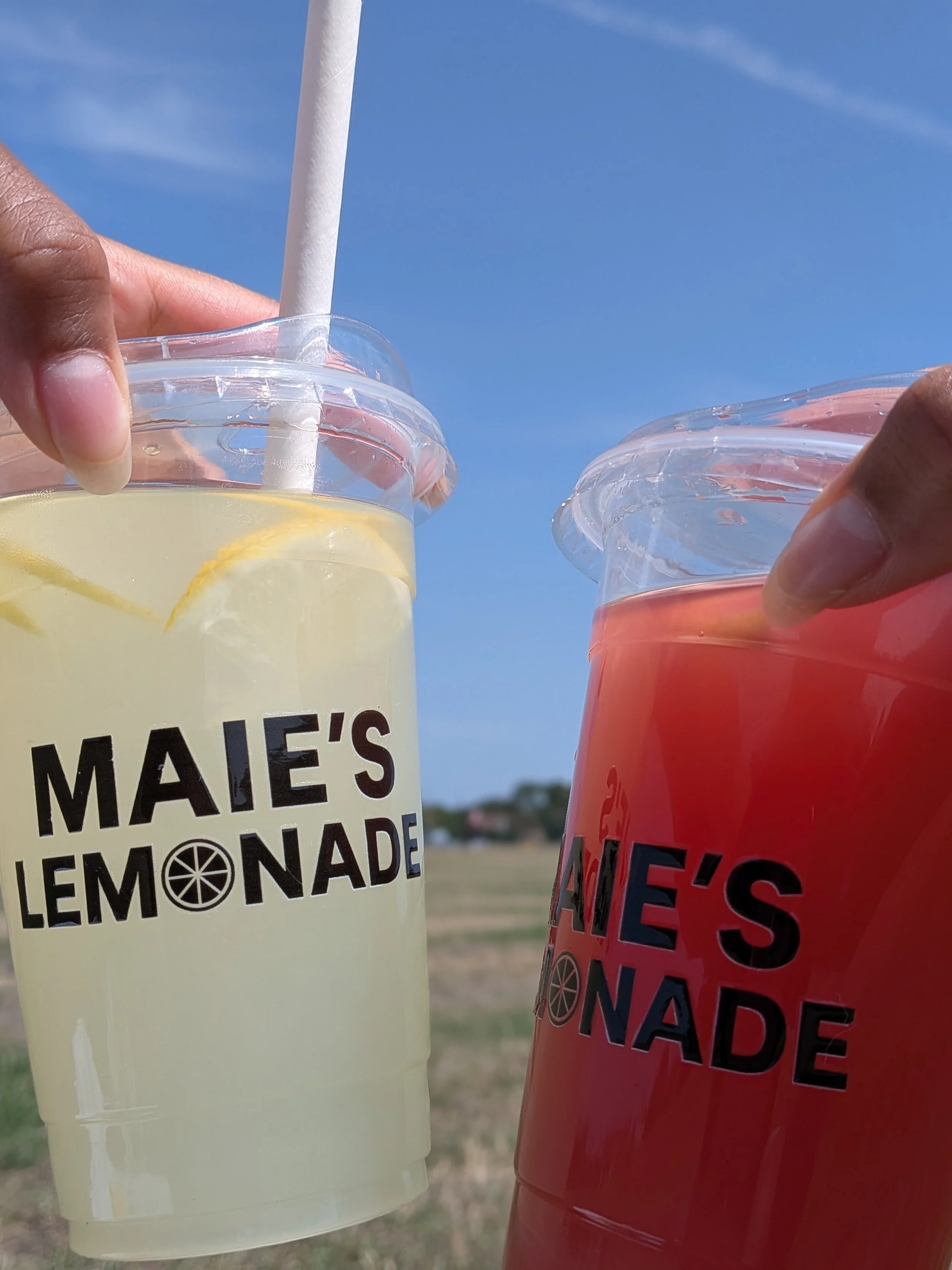Two hands holding cups of lemonade and pink lemonade outdoors, with blue sky and fields in the background.