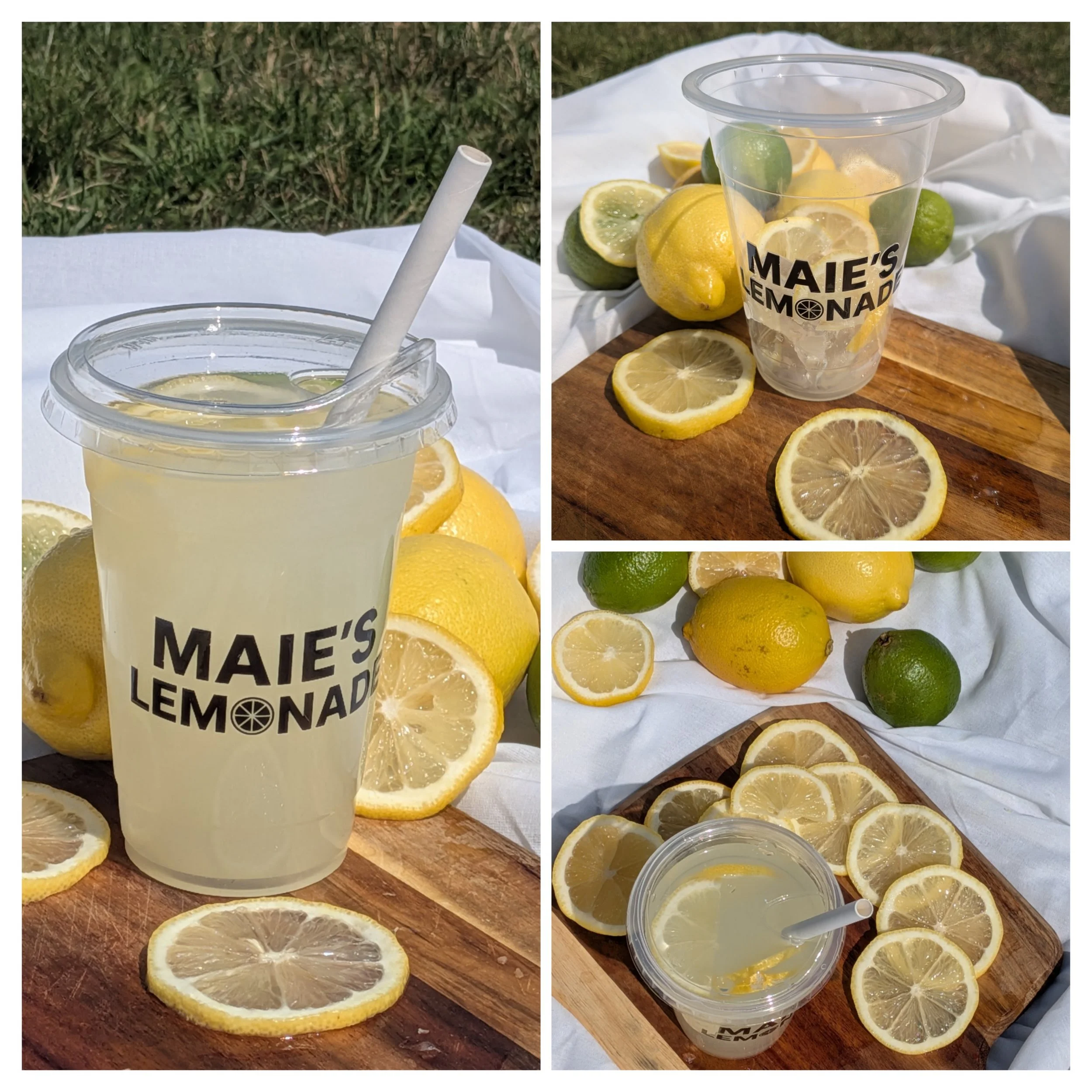 A clear plastic cup labeled "Maie's Lemonade" filled with lemonade, surrounded by fresh lemons and limes, with lemon slices on a wooden cutting board and a white cloth backdrop, outdoors on green grass.