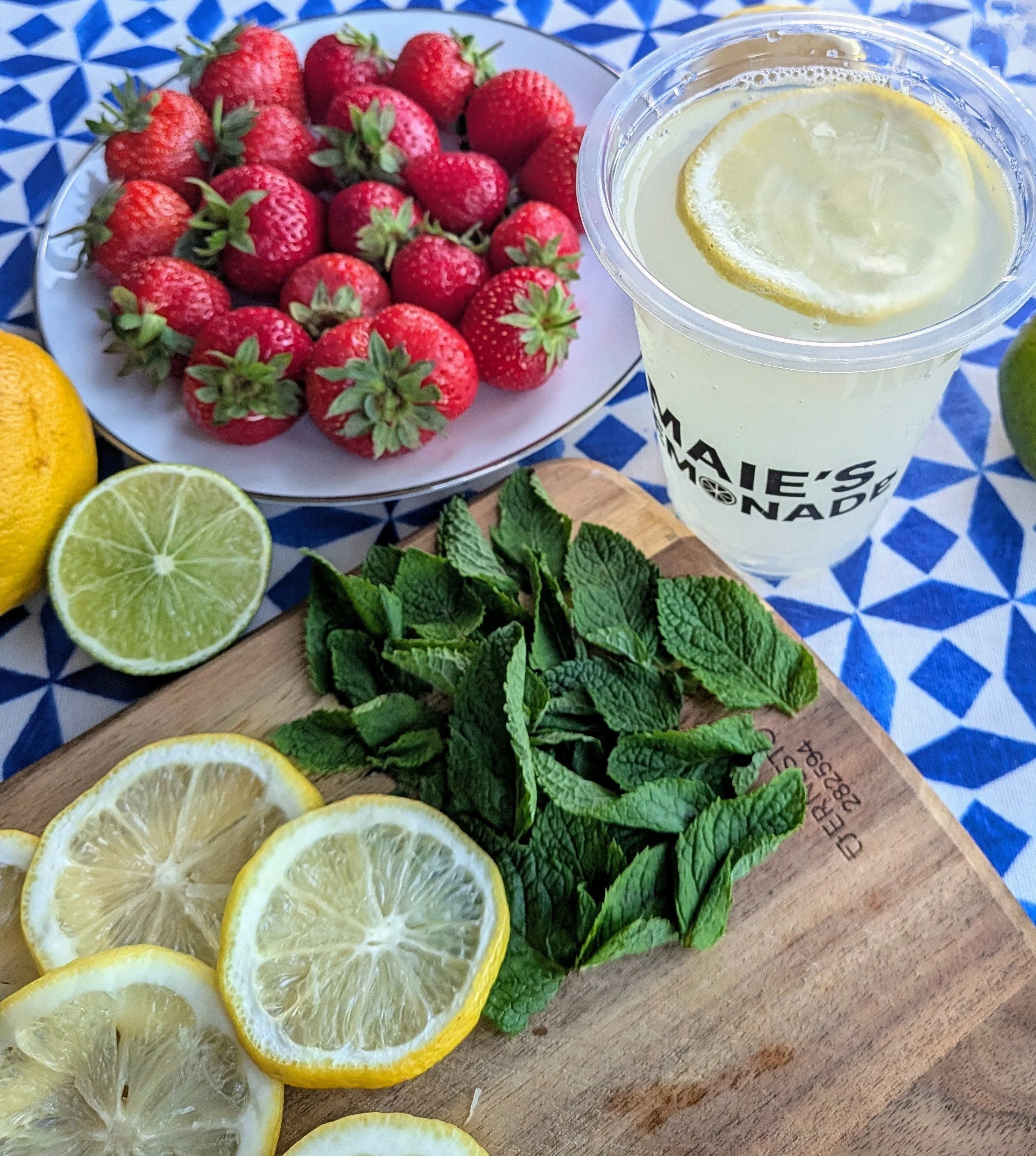 Fresh strawberries on a white plate, sliced lemons and limes, mint leaves on a wooden cutting board, and a cup of lemonade with a lemon slice on top, all placed on a blue and white patterned tablecloth.