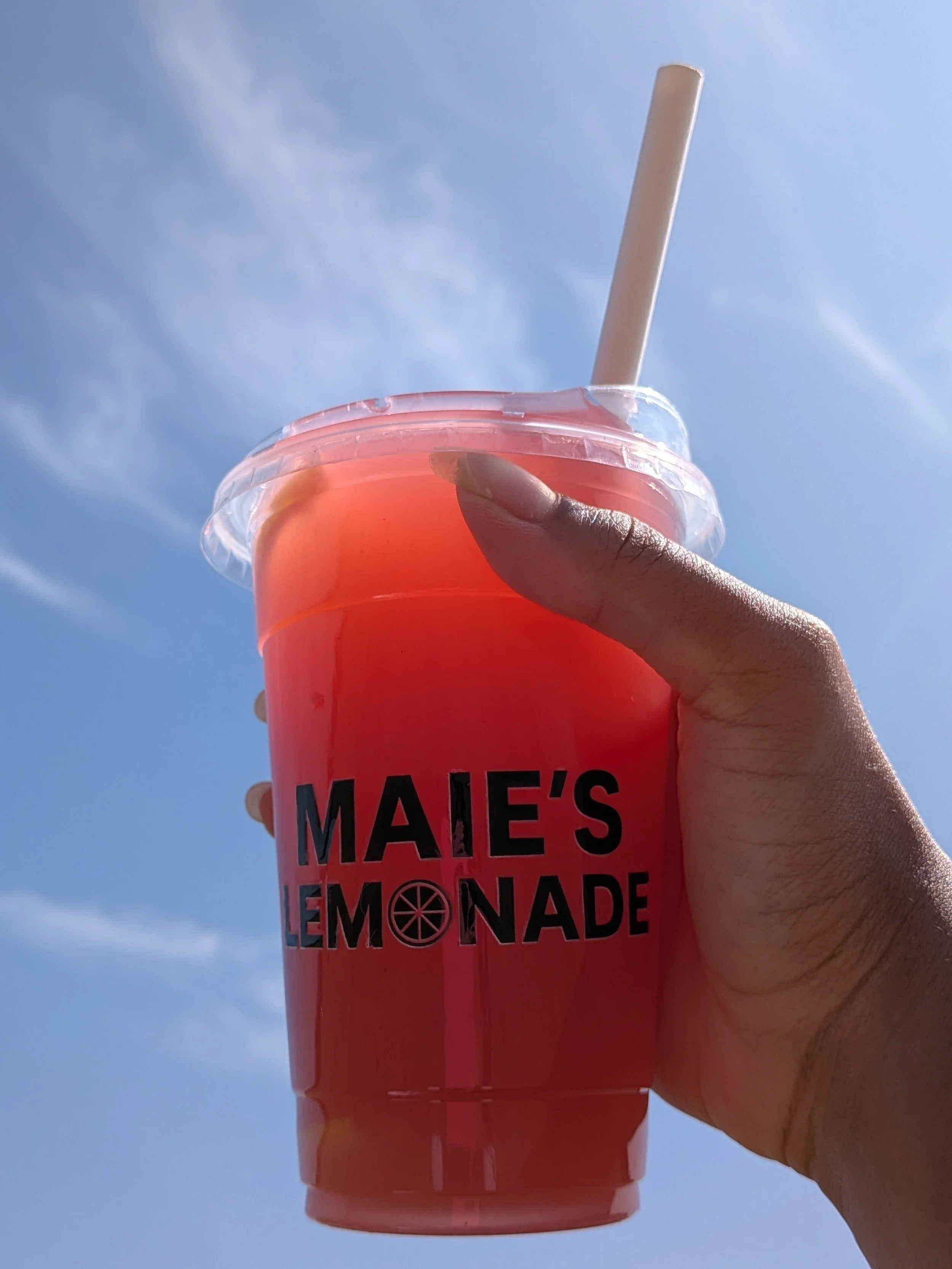 Hand holding a clear plastic cup filled with pink lemonade with a straw, against a bright blue sky with some wispy clouds.