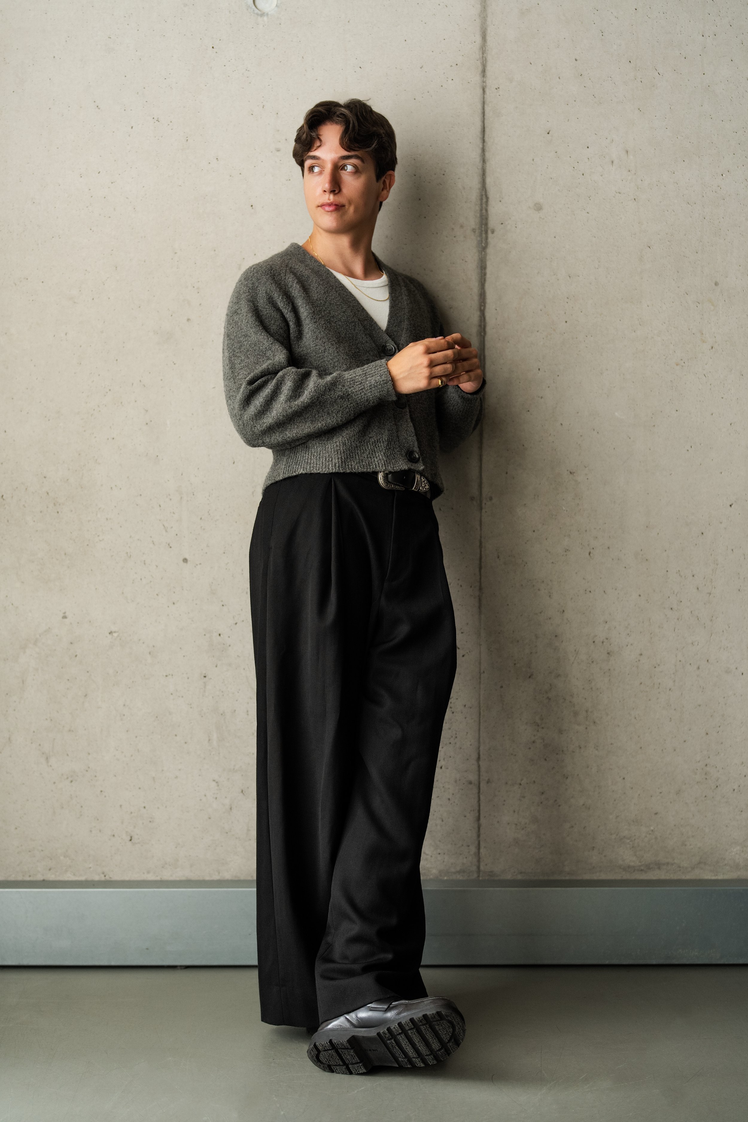 Danny Couto | Couto Studio | wearing a gray cardigan over a white shirt, black high-waisted pants, and loafers, standing against a concrete wall in an indoor setting.