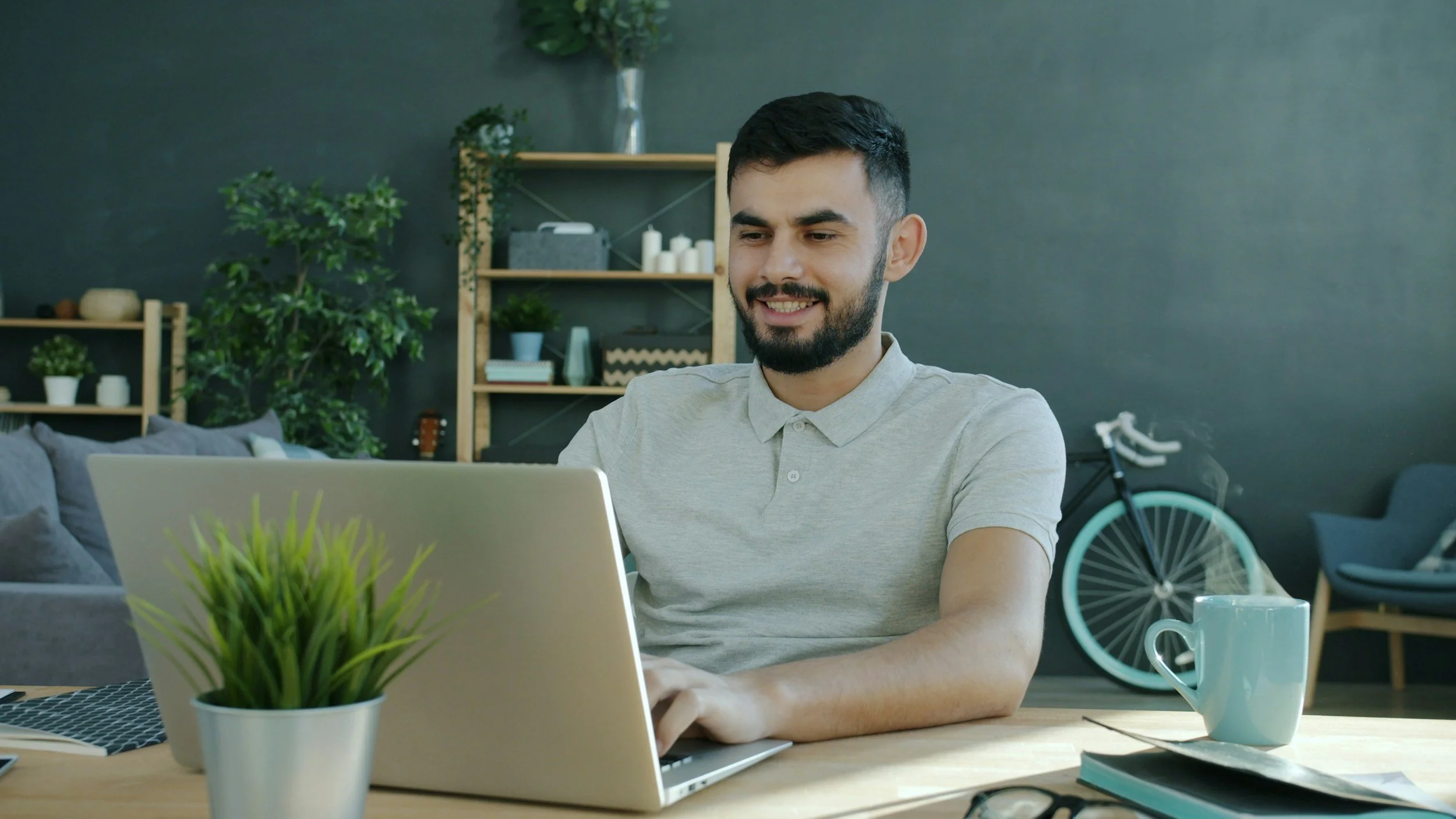 A man smiling and working on a laptop at a wooden table in a cozy living room with bookshelves, plants, a bicycle, and a cup