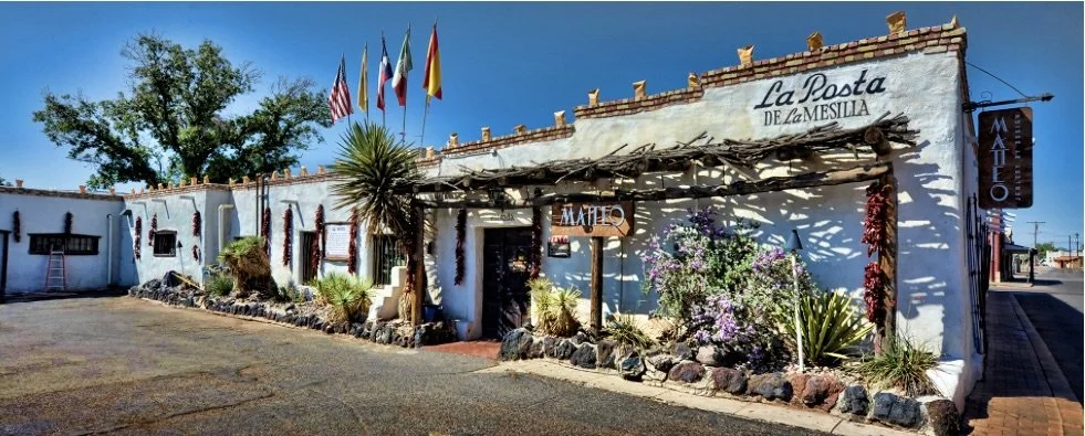A white stucco building with a sign reading 'La Posta de La Mesilla' and 'Matteo' hanging above the entrance, decorated with desert plants, flags, and a wooden pergola exterior, under a bright blue sky.