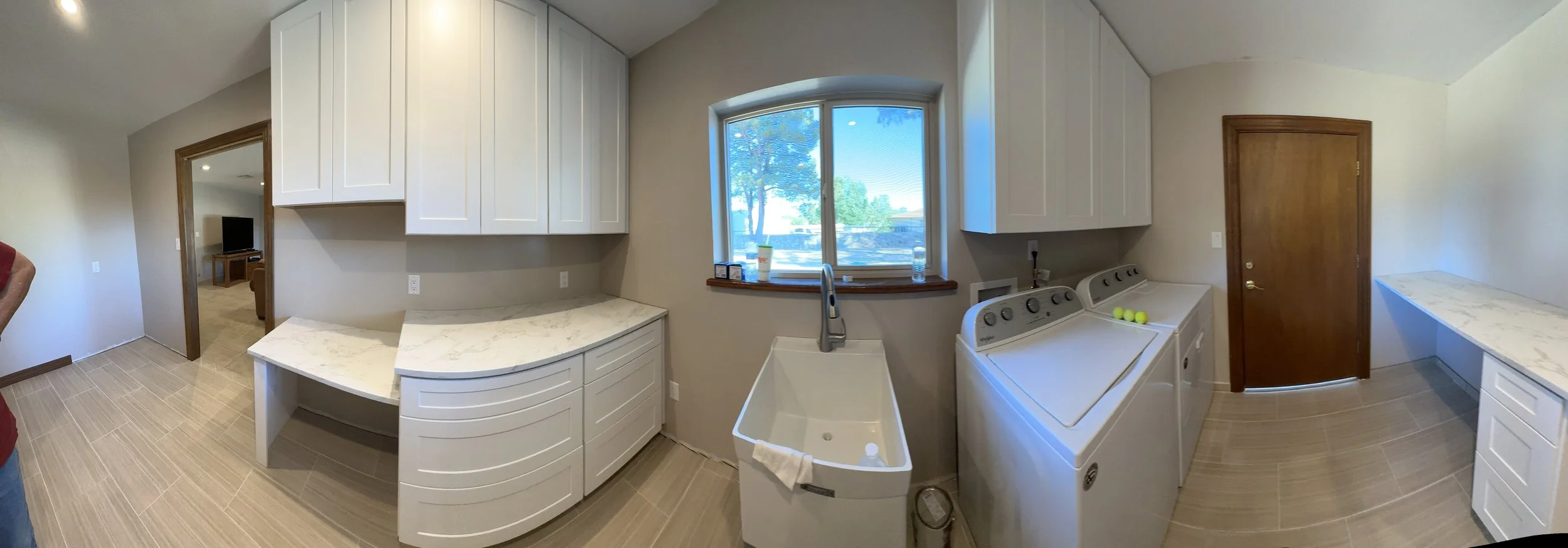 Panoramic view of a laundry room with white cabinets, a large window, a laundry sink, washing machine, and countertop.