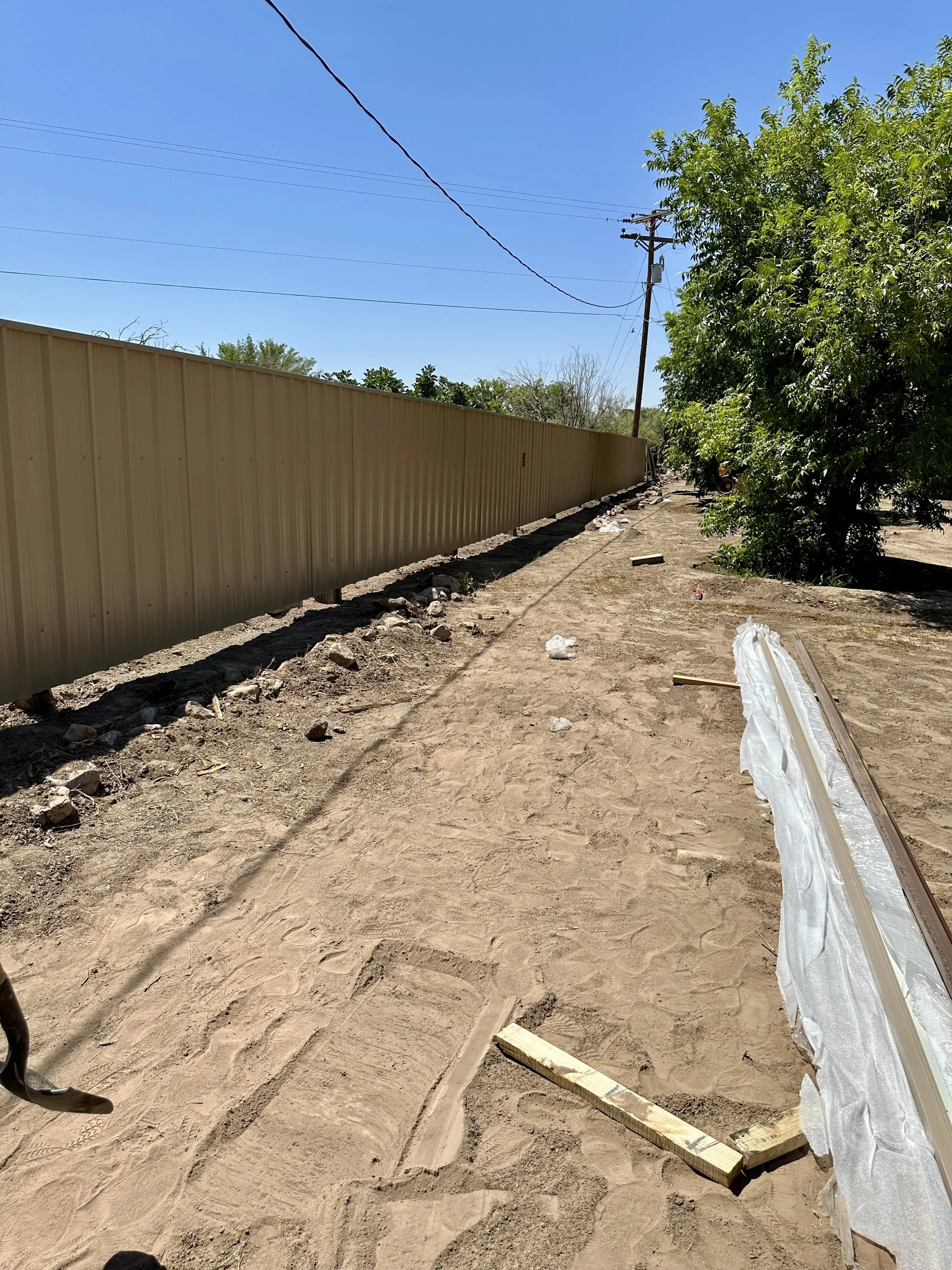 Construction site with a long beige metal fence, dirt ground, scattered debris, and a large leafy tree on the right under a clear blue sky.