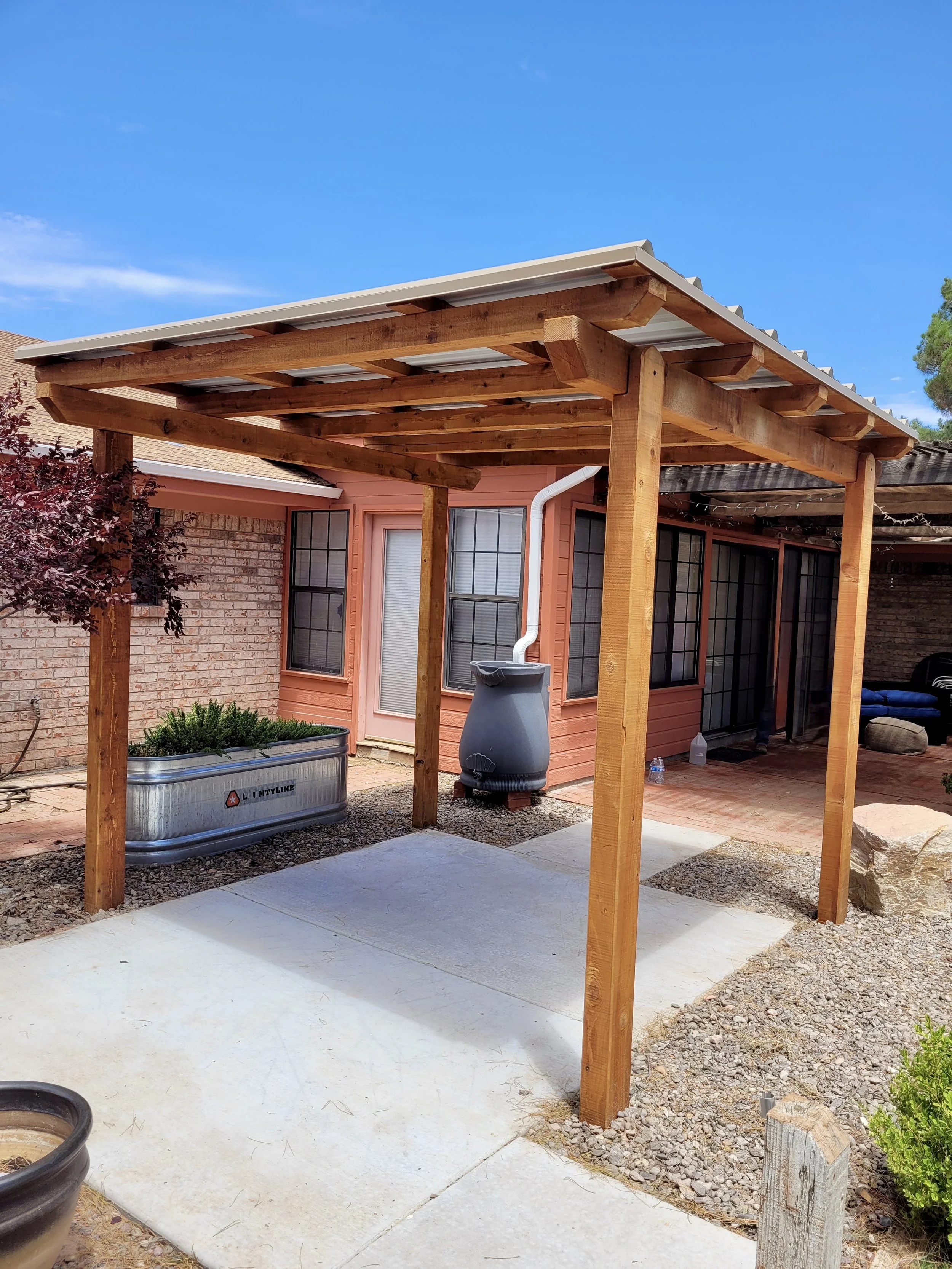 Backyard patio with a newly built wooden pergola, attached to a brick house with pink siding, multiple windows, a rain barrel, and a concrete patio area.