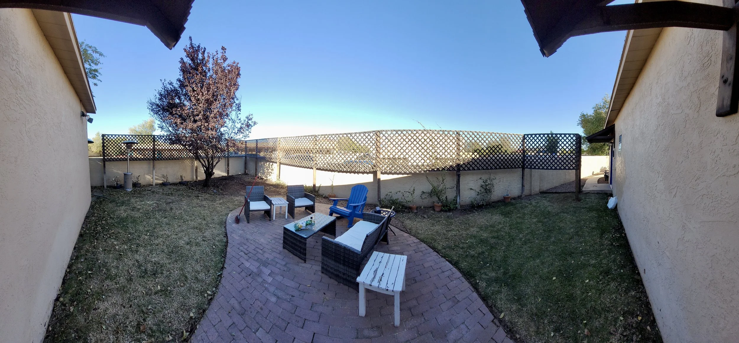 Backyard patio with brick flooring, patio furniture including chairs and a table, a tree with reddish leaves, and a wooden lattice fence under a clear blue sky.