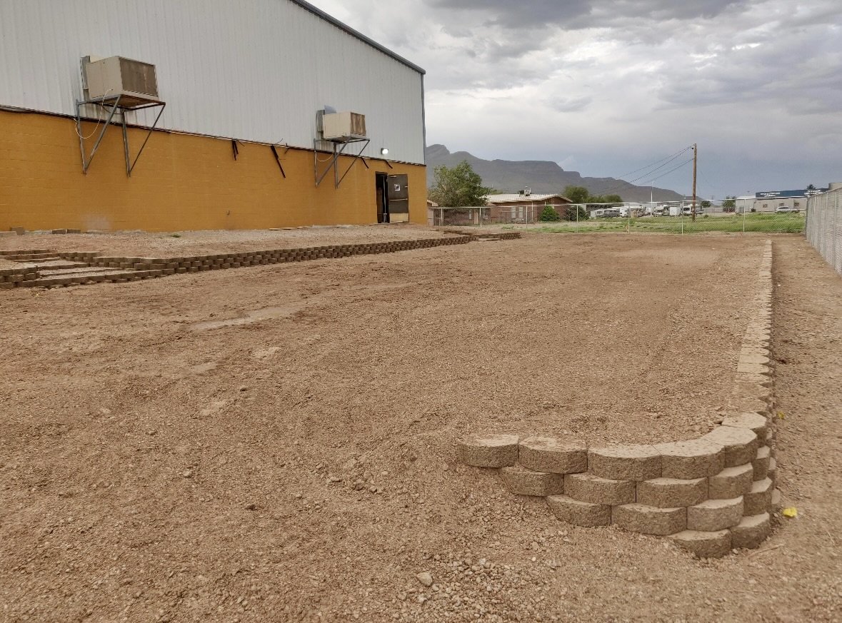A dirt lot with a partially built brick wall and steps near a large industrial building, with mountains and cloudy sky in the background.