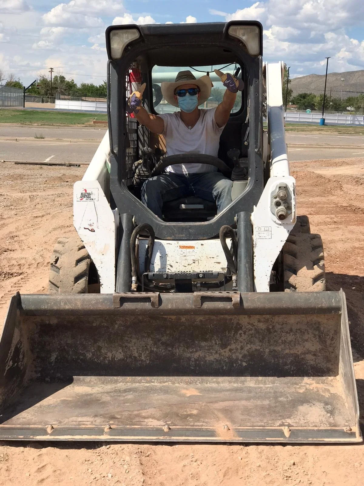 Person sitting inside a small construction vehicle, wearing a cowboy hat, sunglasses, face mask, and gloves, on a dirt construction site.