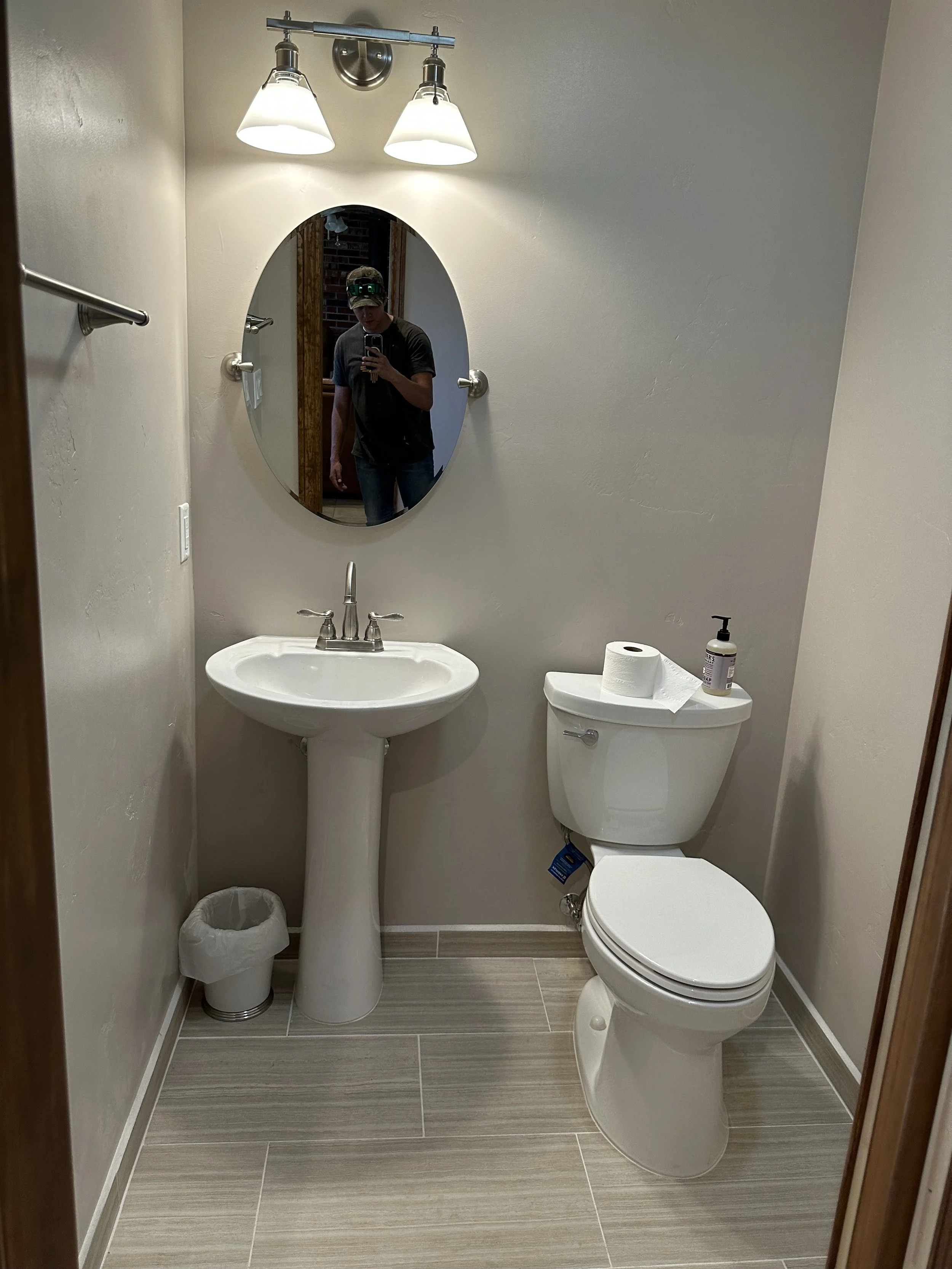 Small bathroom with a pedestal sink, a toilet with a roll of toilet paper and soap on top, a mirror, and a wall-mounted light fixture. The floor is tiled with beige horizontal stripes, and there's a person taking a selfie in the mirror.