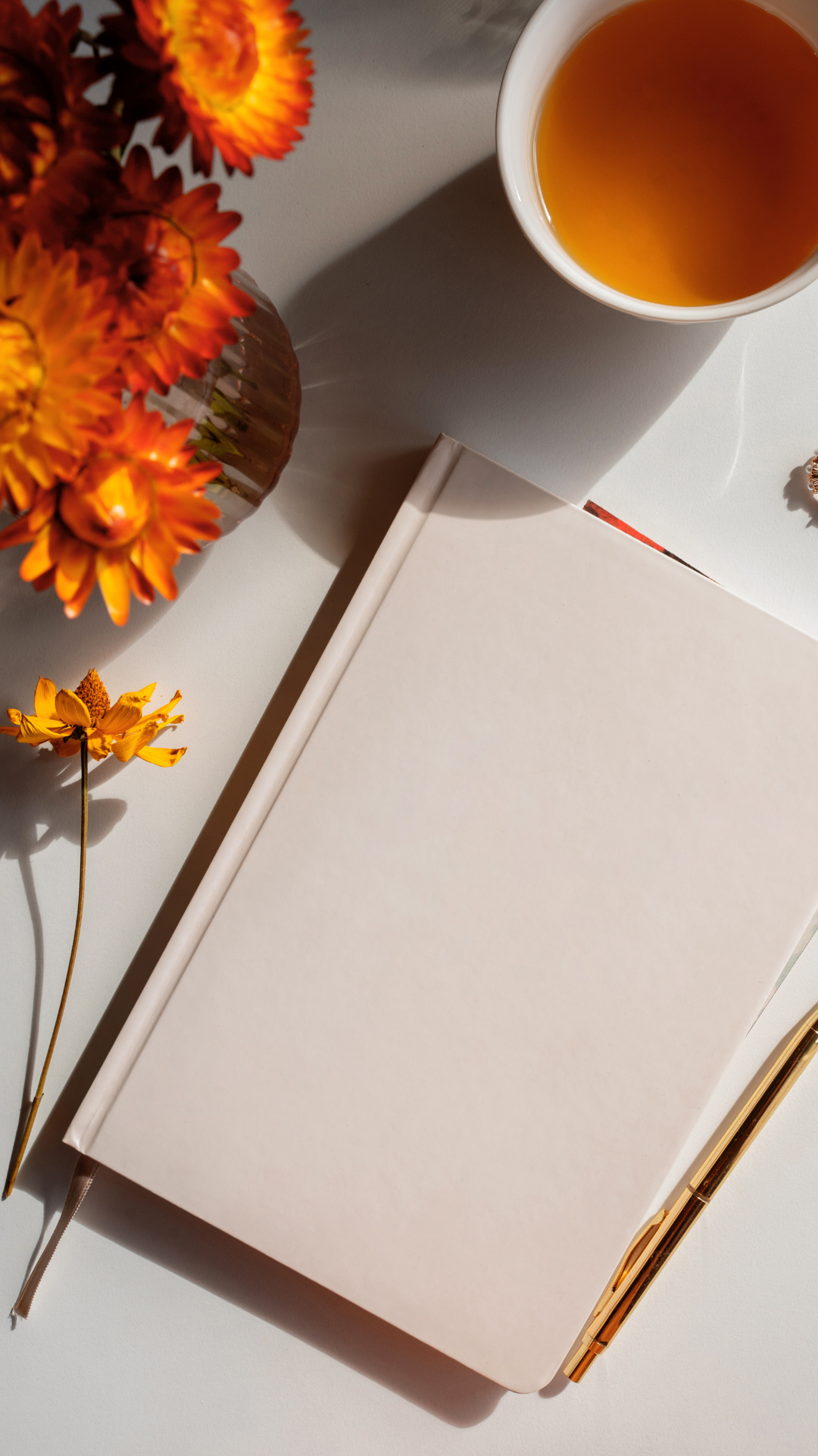 An overhead view of a white closed notebook with a gold pen resting on it. To the left, there is a small bouquet of orange flowers in a vase. To the upper right, there is a white cup filled with tea, casting a shadow onto the white surface.