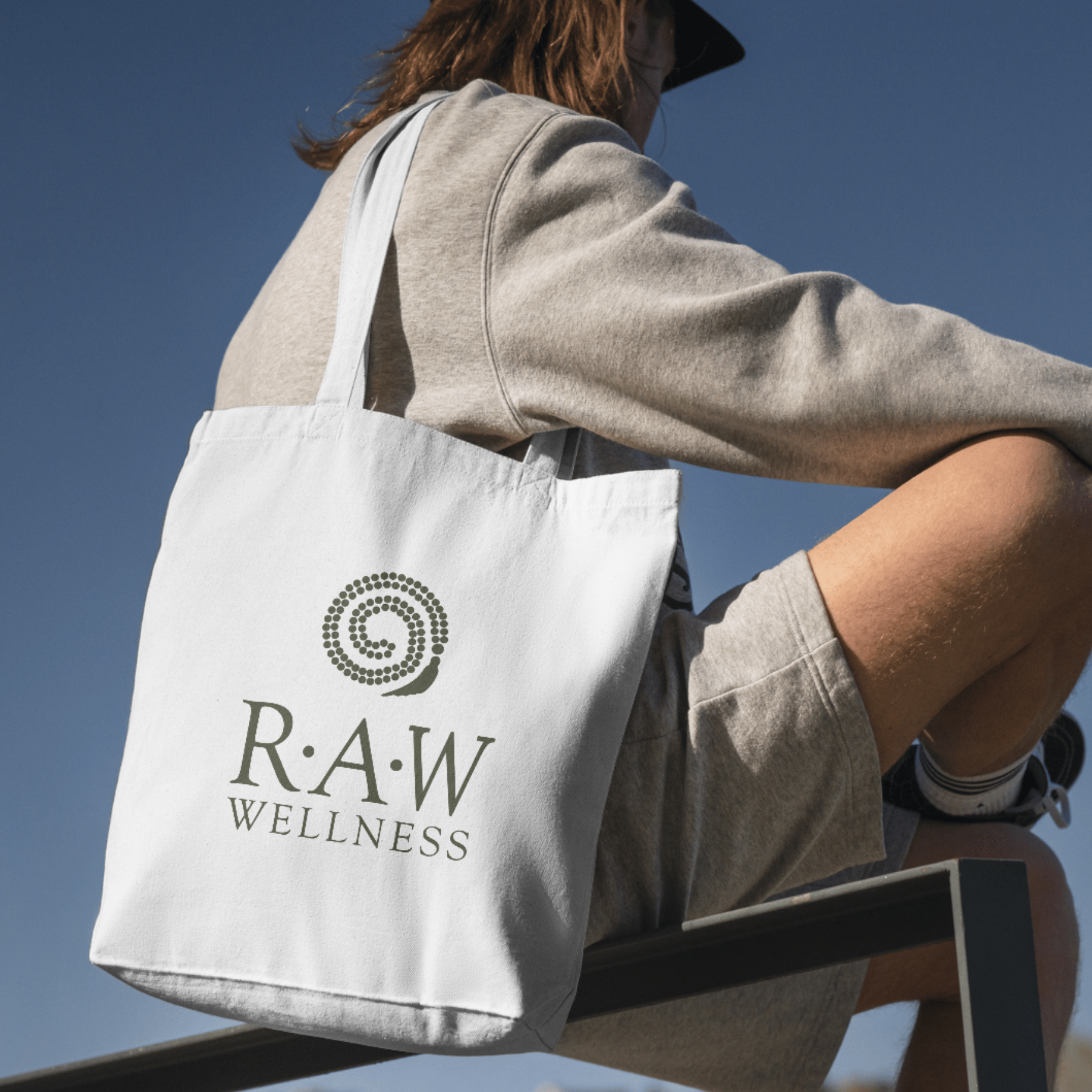 A person wearing a gray sweatshirt and shorts, sitting outdoors against a clear blue sky, carrying a white tote bag with the logo and text 'R.A.W WELLNESS' printed on it.