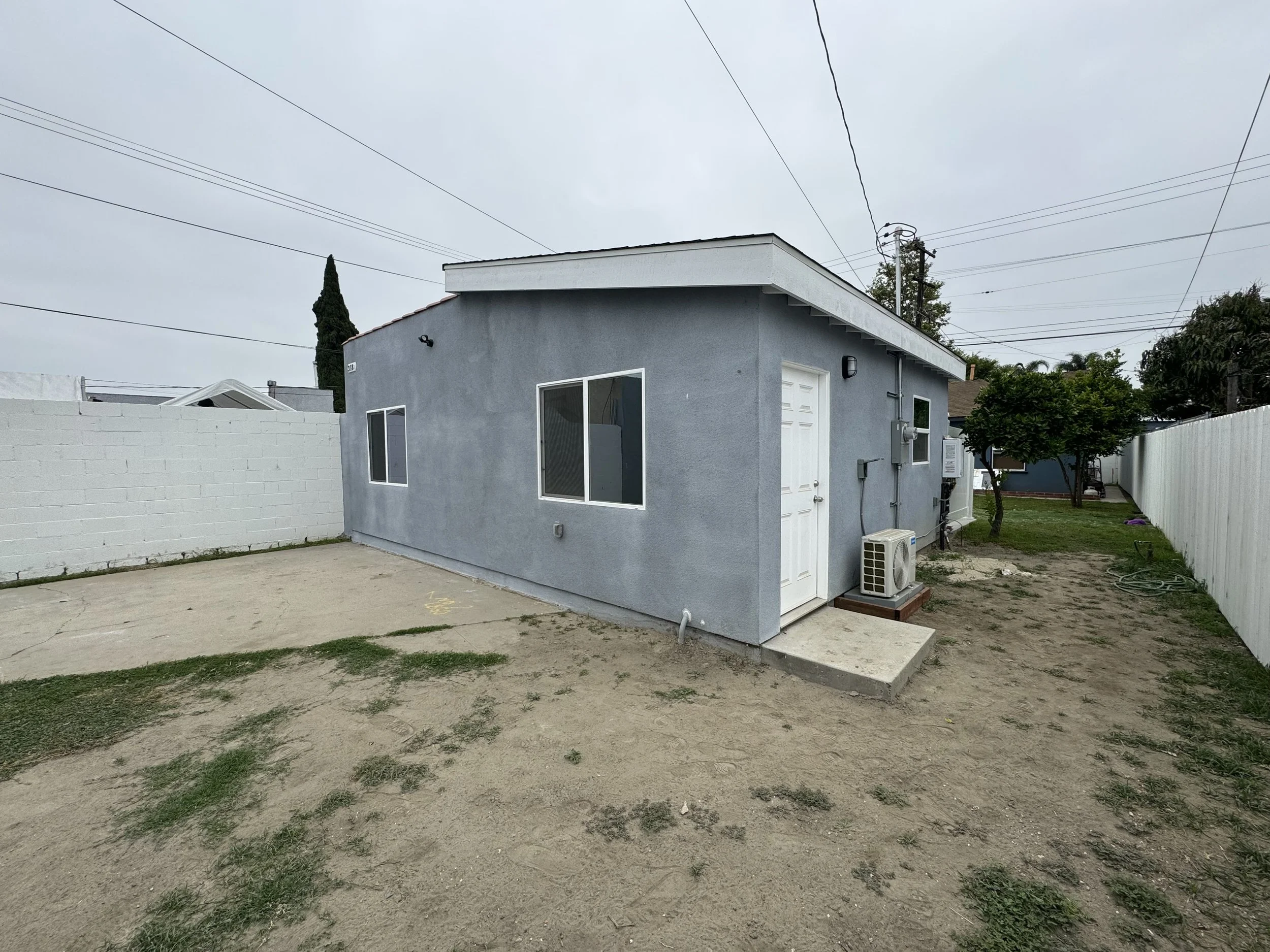 Small gray house with white door and three windows, air conditioning unit outside, on a partly grassy yard with a dirt area and a concrete patio, enclosed by a white fence on one side and a white wall on the other, overcast sky.