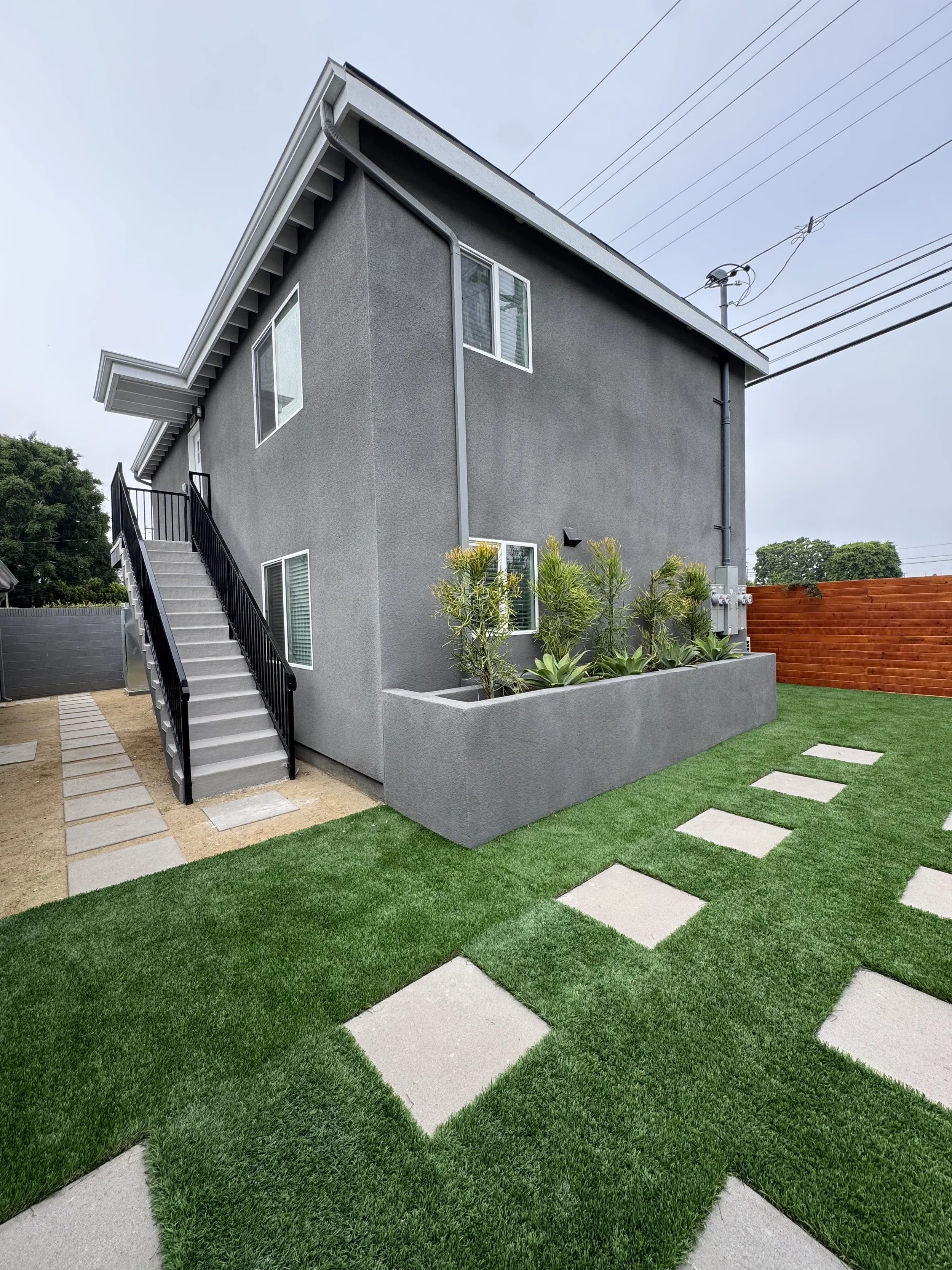 A modern gray two-story house with a staircase on the side, surrounded by a green lawn with concrete stepping stones, a raised flower bed with plants, and a wooden fence in the backyard.