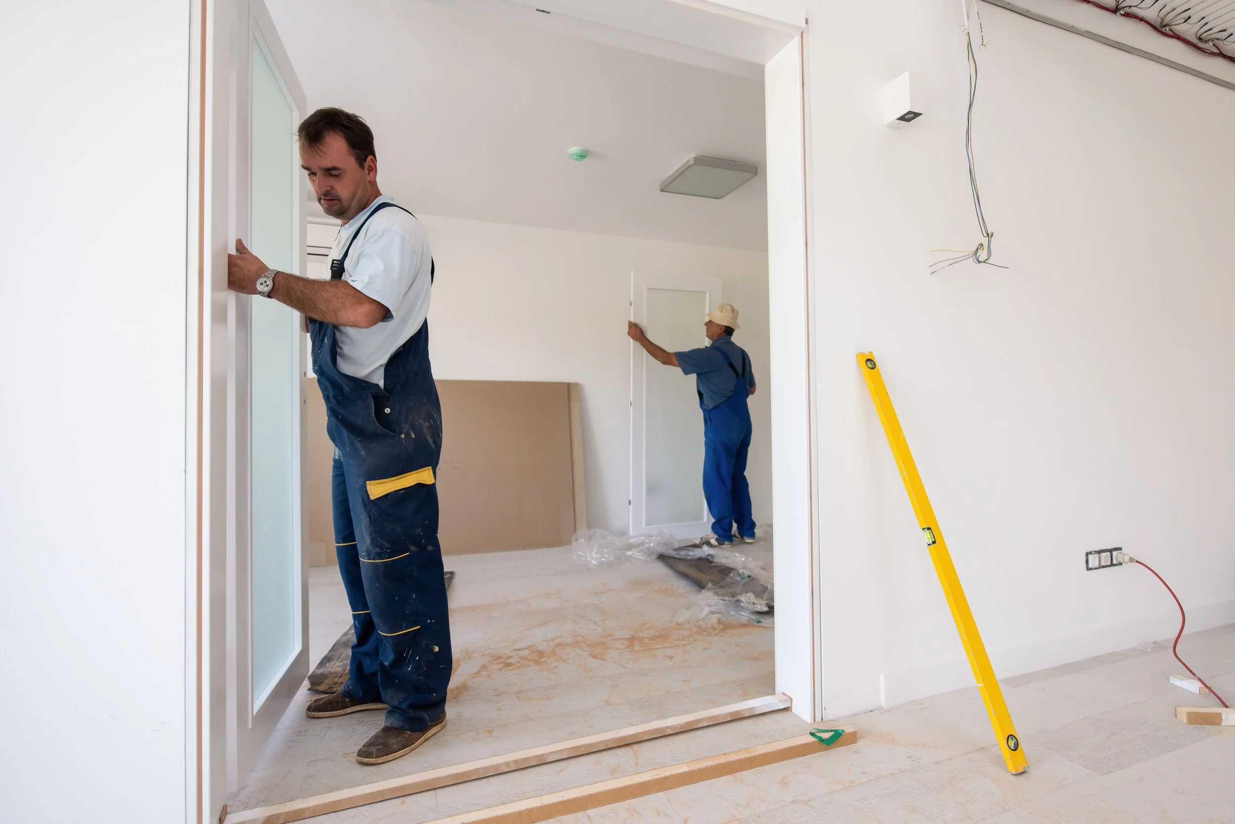 Two men installing a door inside a new home under construction. One man is holding a frosted glass panel, and the other is adjusting the door frame in the background.