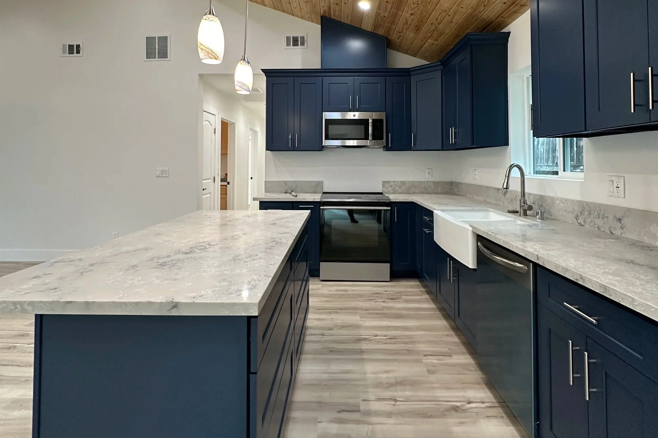 Modern kitchen with dark blue cabinets, white and gray marble countertops, a large kitchen island, stainless steel appliances including microwave and oven/stove, white farmhouse sink, wooden ceiling, and pendant lighting.