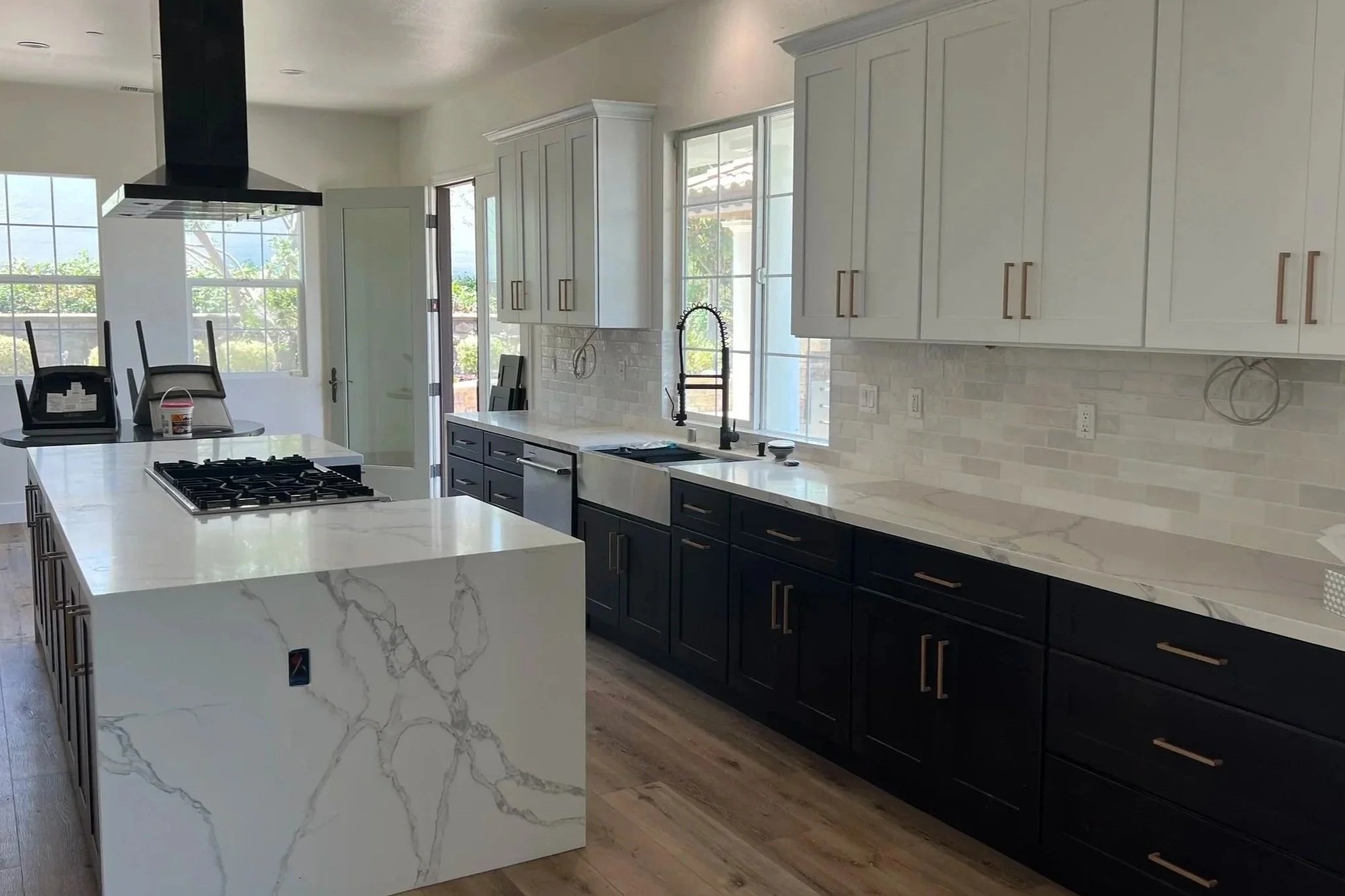 Modern kitchen with white and black cabinetry, marble countertops, and large windows letting in natural light. An island with a marble surface and cooking stove is in the foreground, and a black range hood hangs overhead.