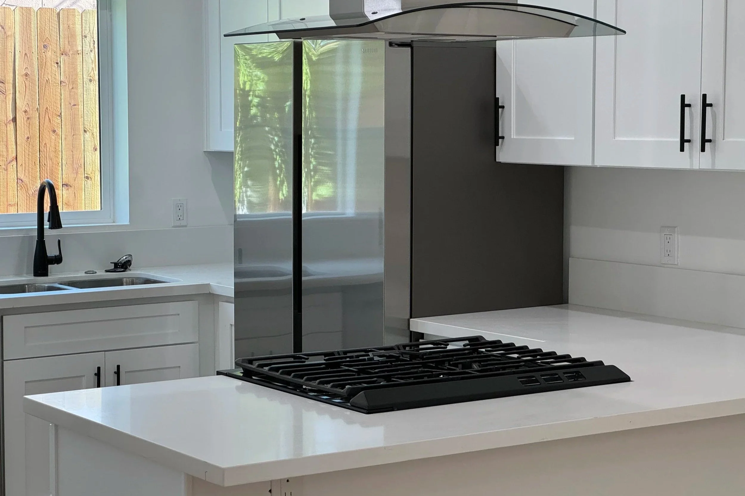 Modern kitchen with white cabinets, a white countertop, a black faucet, a stainless steel refrigerator, black handles on the cabinets, and a black gas stovetop, with a window showing a wooden fence outside.