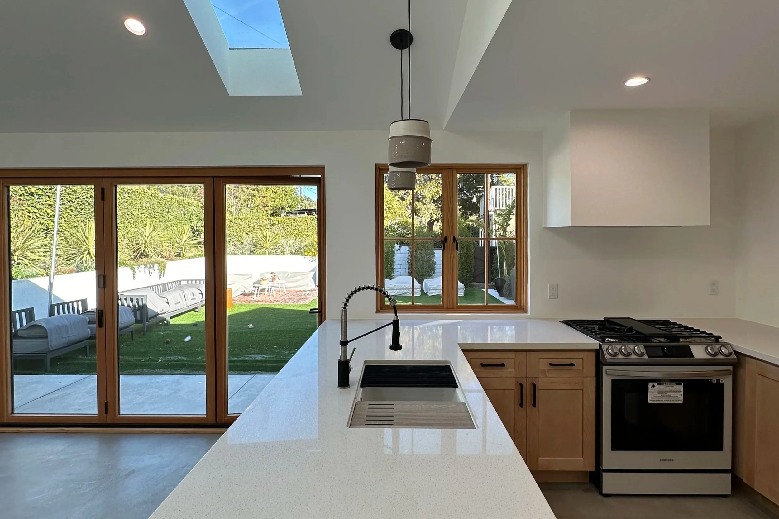 Modern kitchen with white countertops, wooden cabinets, stainless steel stove, black sink faucet, and large windows and sliding doors opening to a backyard with outdoor seating.