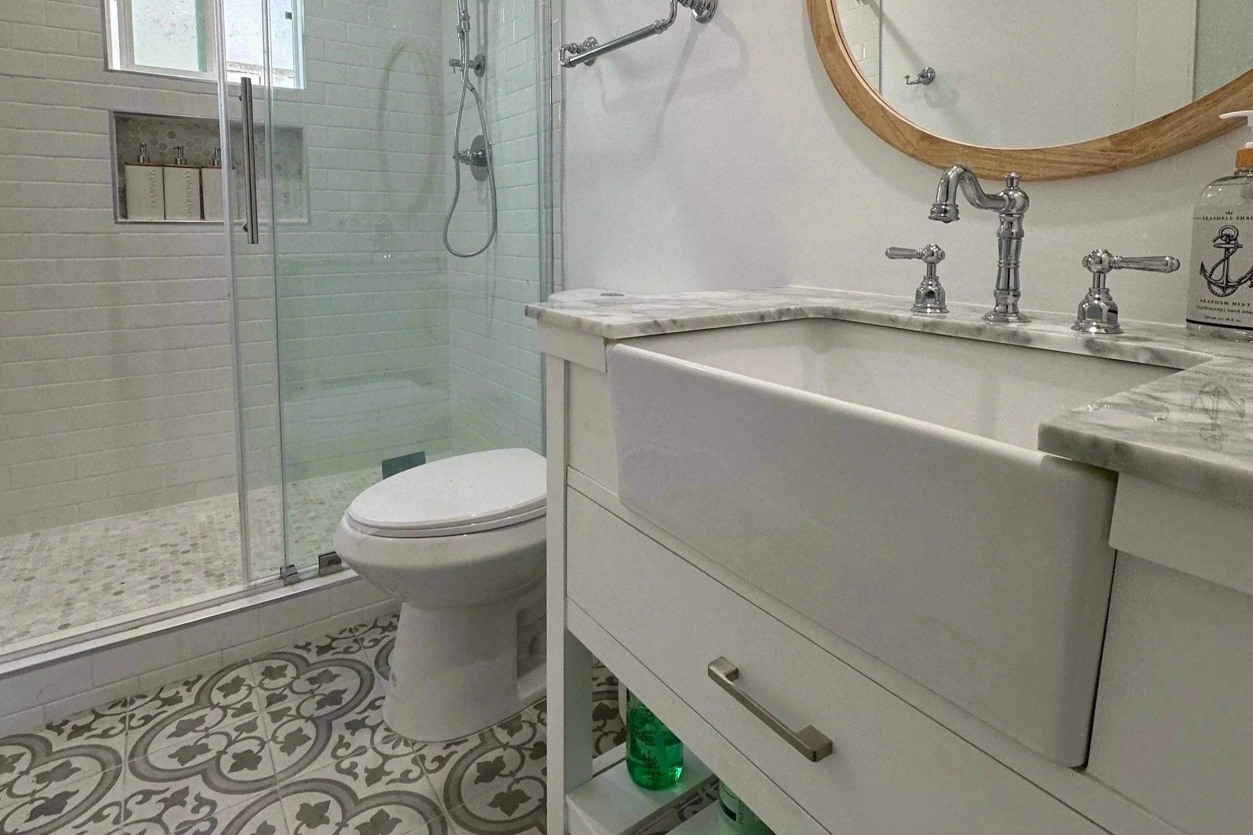 A modern bathroom with a glass-enclosed shower, white brick wall tiles, a toilet, white vanity with a marble countertop, and a round wooden mirror above the sink.