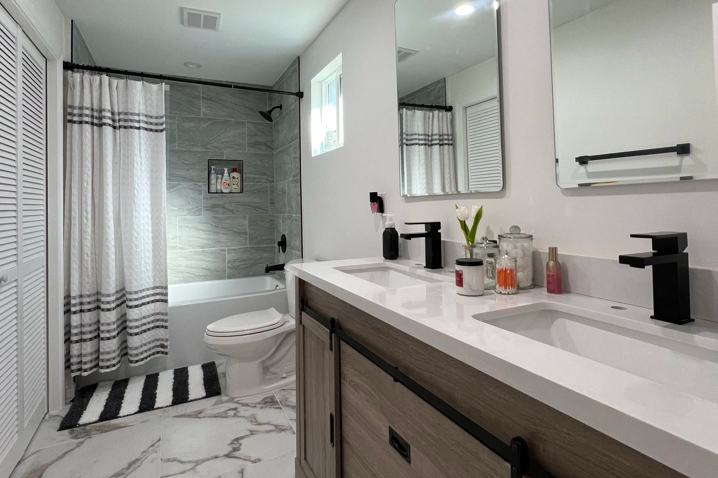 Modern bathroom with dual sinks, black faucets, large mirror, window, bathtub with shower, gray tile walls, striped shower curtain, white and gray marble floor, decorative jars, and small flower on countertop.