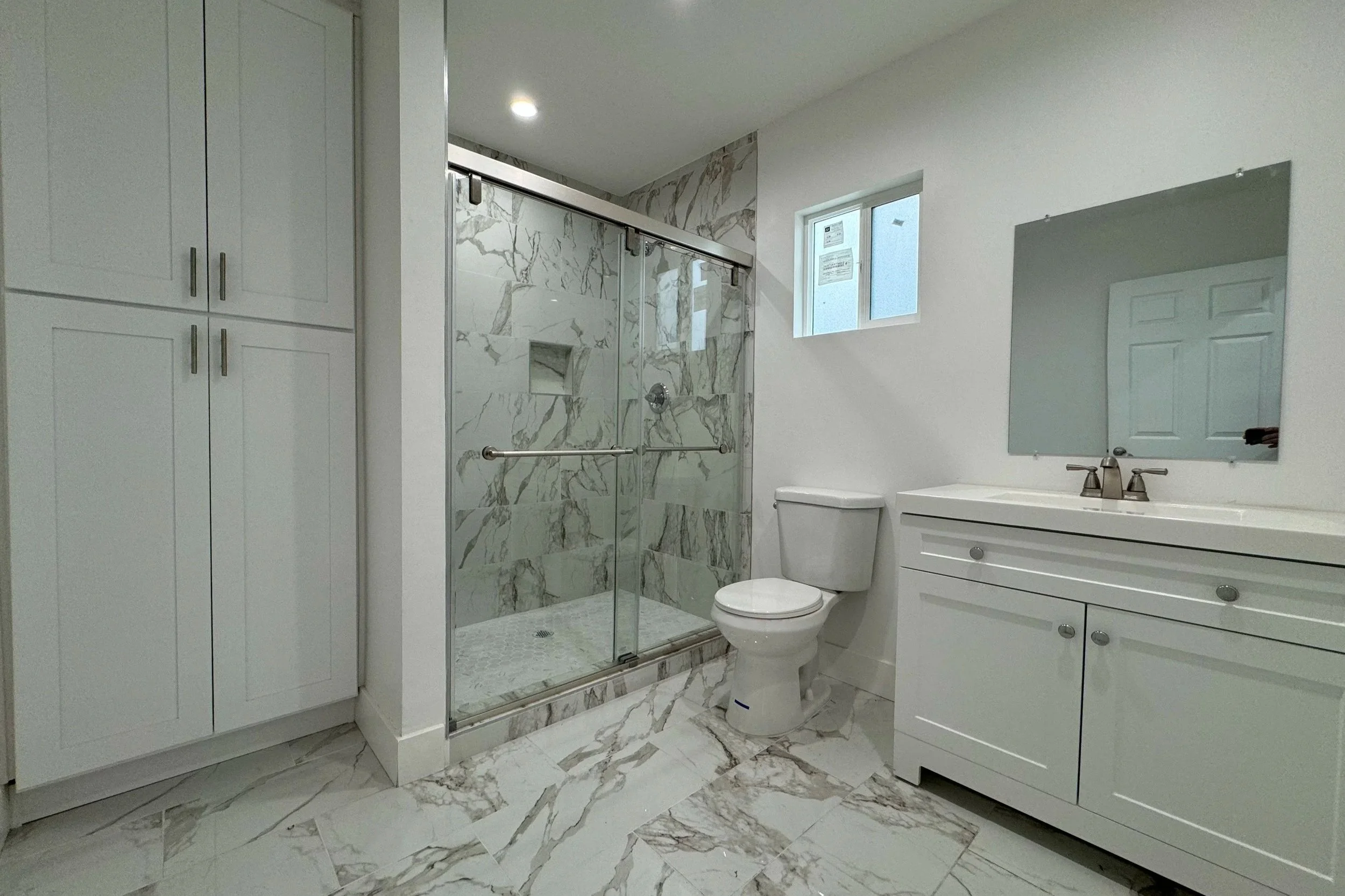 Modern bathroom with marble floors and shower, white cabinetry, toilet, and rectangular mirror