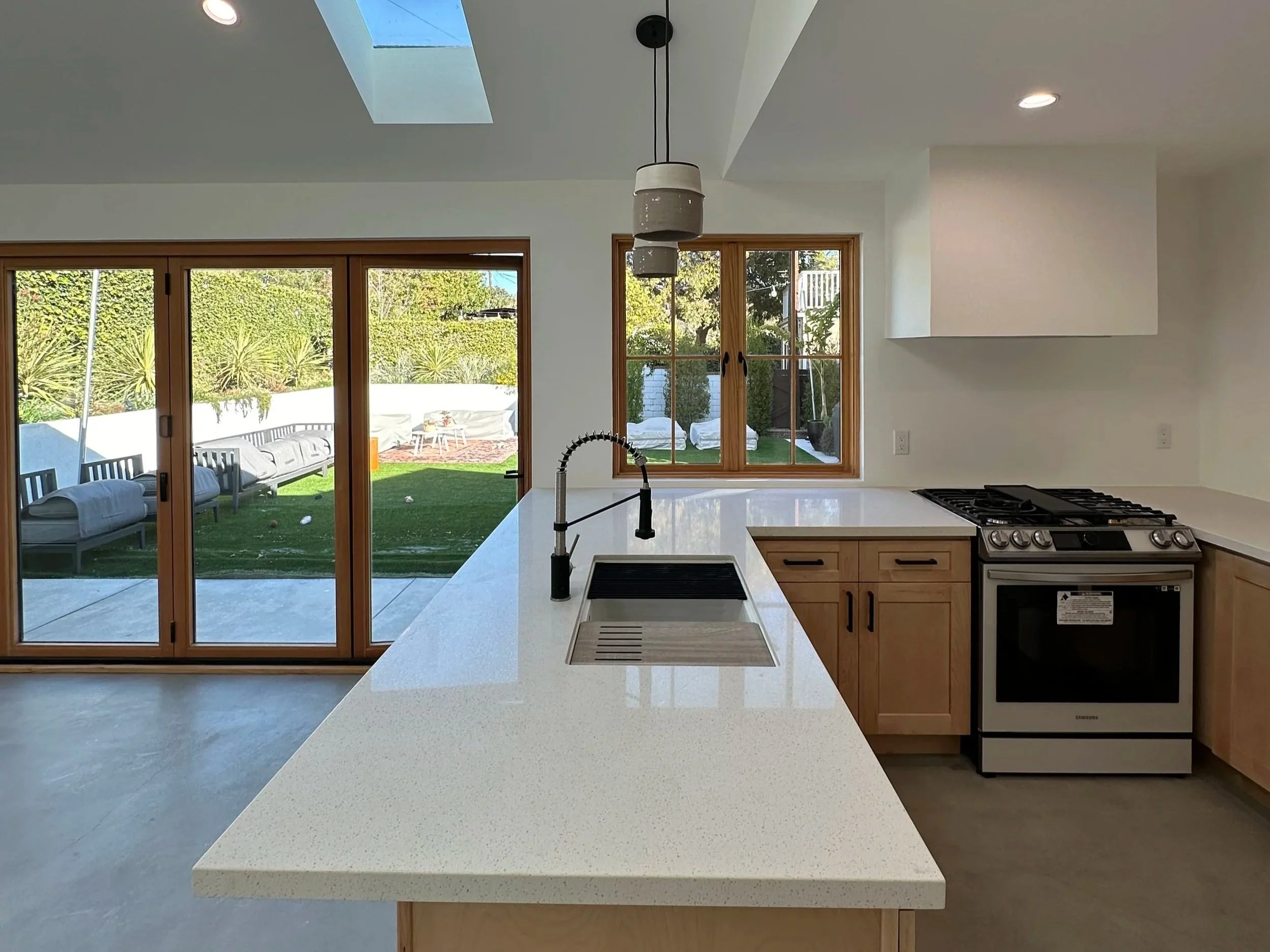Modern kitchen with white countertops, wooden cabinets, a stainless steel stove, large window, sliding glass doors opening to a backyard patio with outdoor furniture and greenery, and pendant lights hanging above the island.