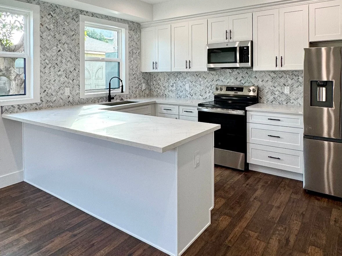 Modern kitchen with white cabinets, stainless steel appliances, a marble backsplash, dark hardwood floors, and two large windows.