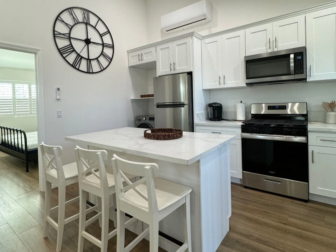 Modern kitchen with white cabinets, stainless steel refrigerator, microwave, oven, and a small kitchen island with three white chairs. There is a large clock on the wall and an air conditioning unit.
