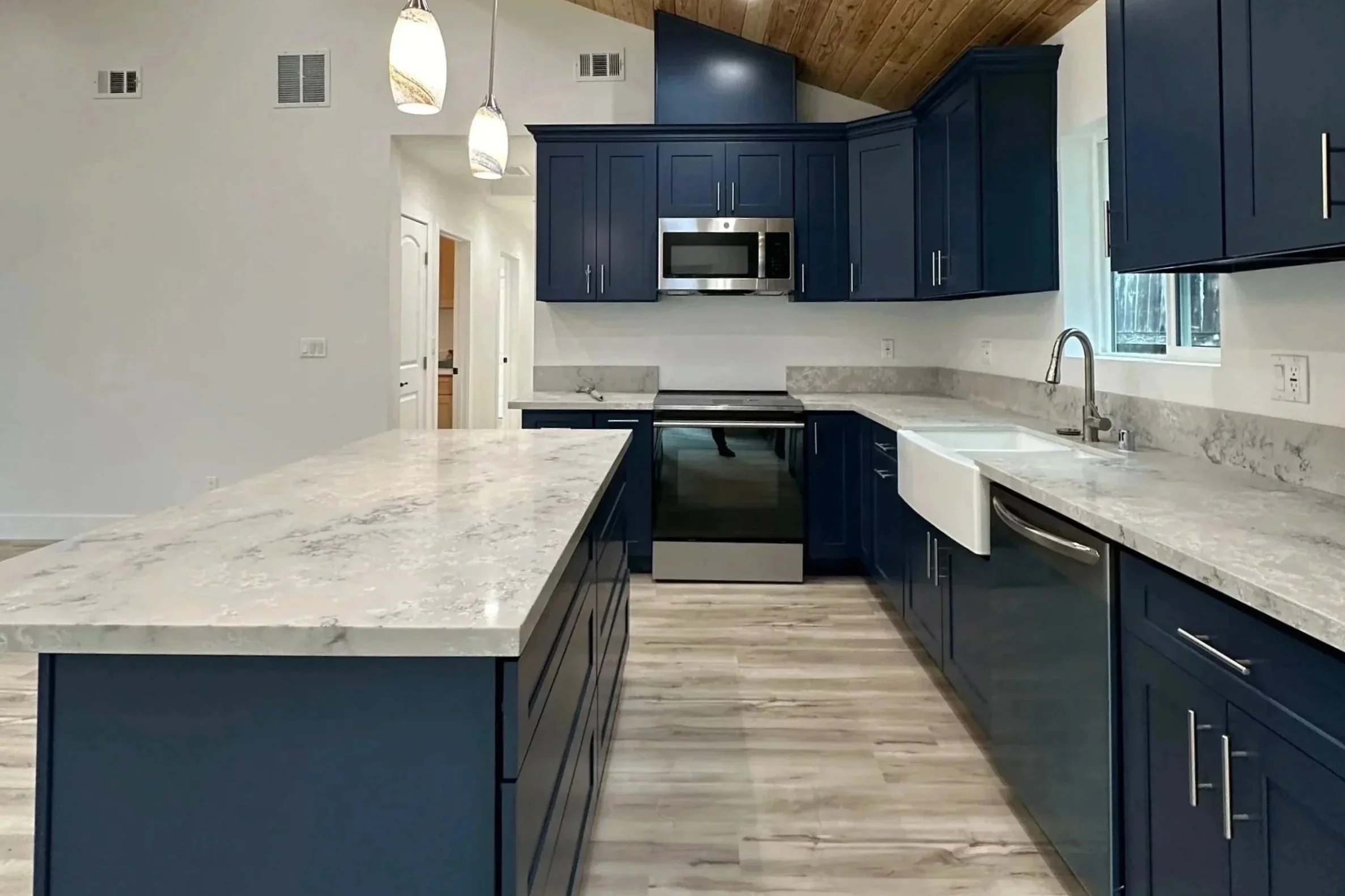 Modern kitchen with blue cabinets, marble countertops, a white farmhouse sink, stainless steel microwave and stove, light wood flooring, and pendant lighting.