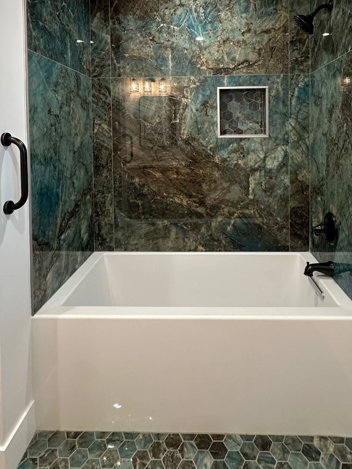 Bathroom shower with green and brown marble tiles, built-in niche, black fixtures, and a white bathtub.