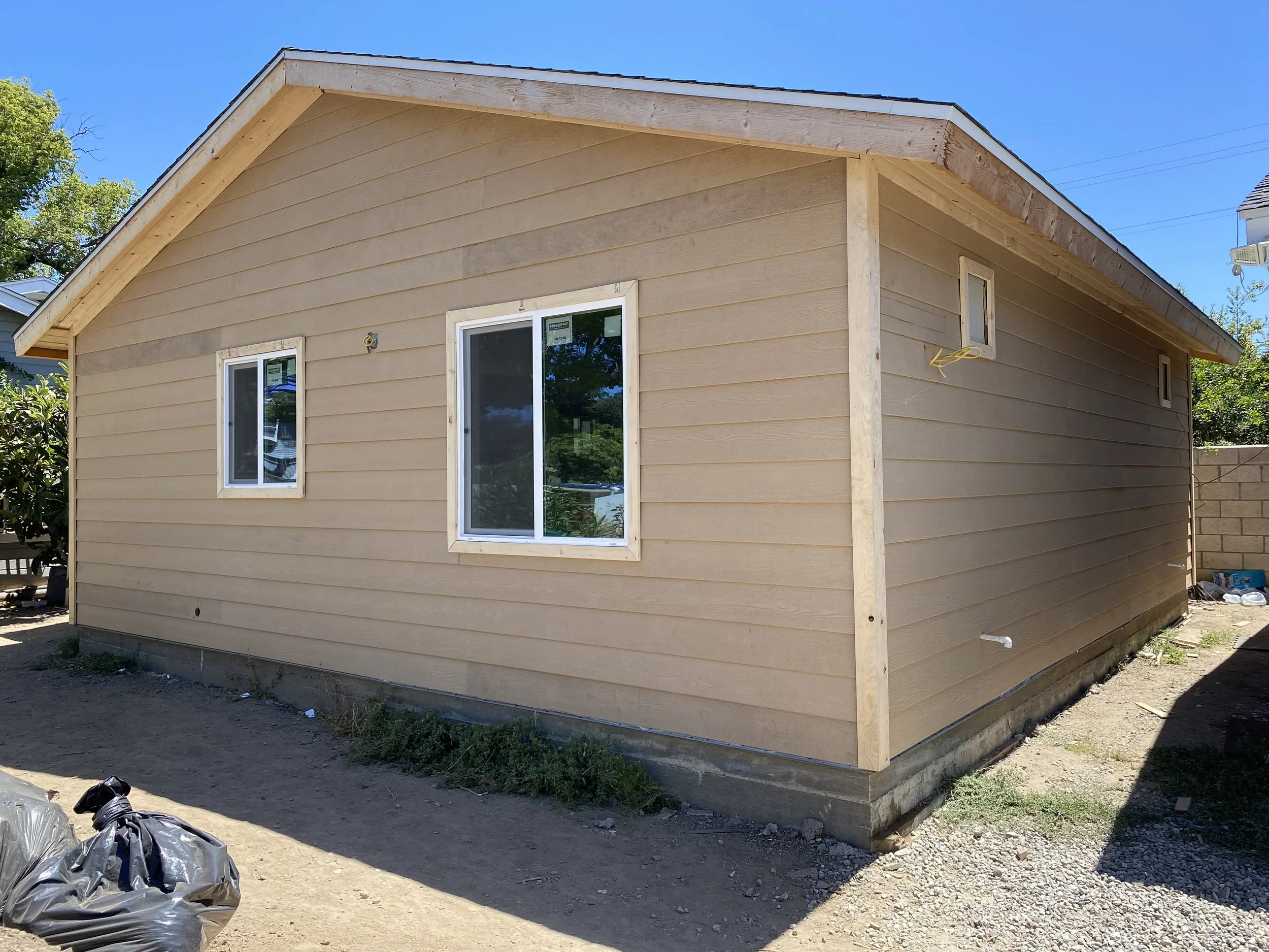 Side view of a house with beige siding and white-framed windows under a blue sky, with some construction materials and debris nearby.