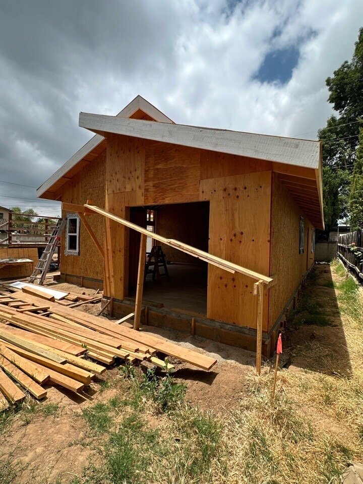 A house under construction with wooden exterior walls, a sloped roof, and construction materials and tools in the foreground.