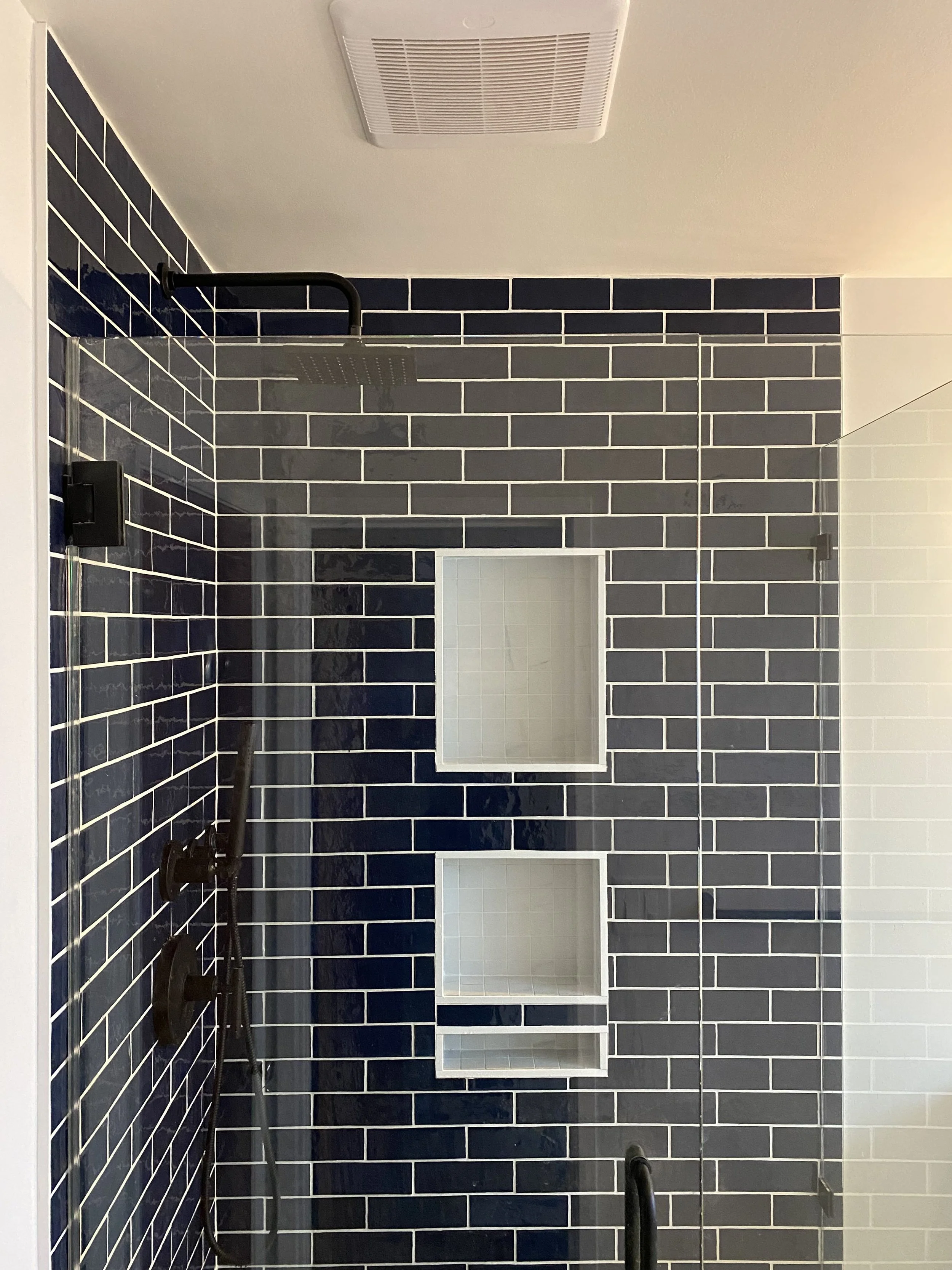 Bathroom shower with black subway tile walls, a large rainfall showerhead mounted on a black pipe, a small built-in niche with three shelves, and a clear glass shower door.
