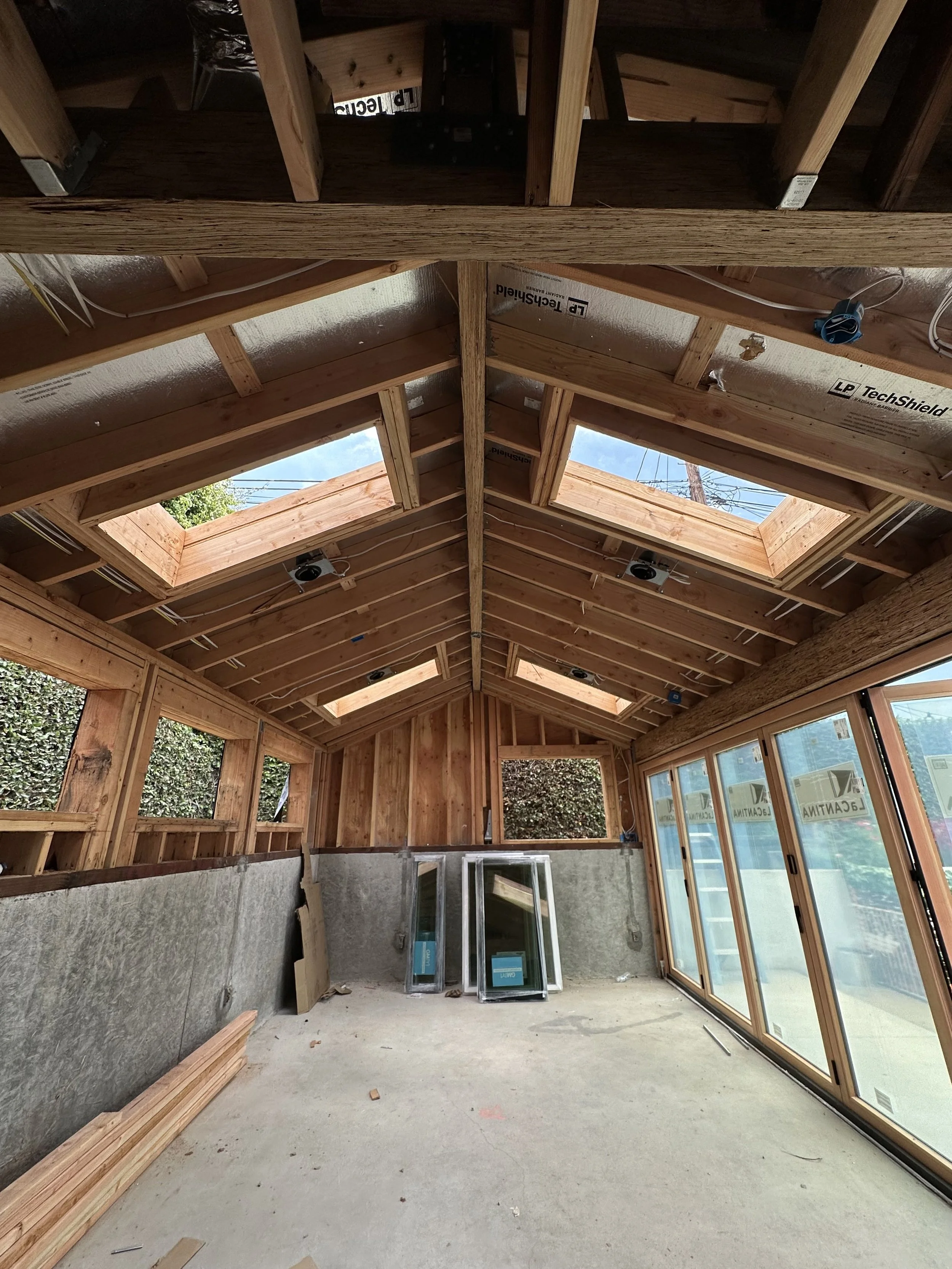 The interior of a house under construction showing exposed wooden beams, framing, skylights in the roof, and sliding glass doors along one wall.