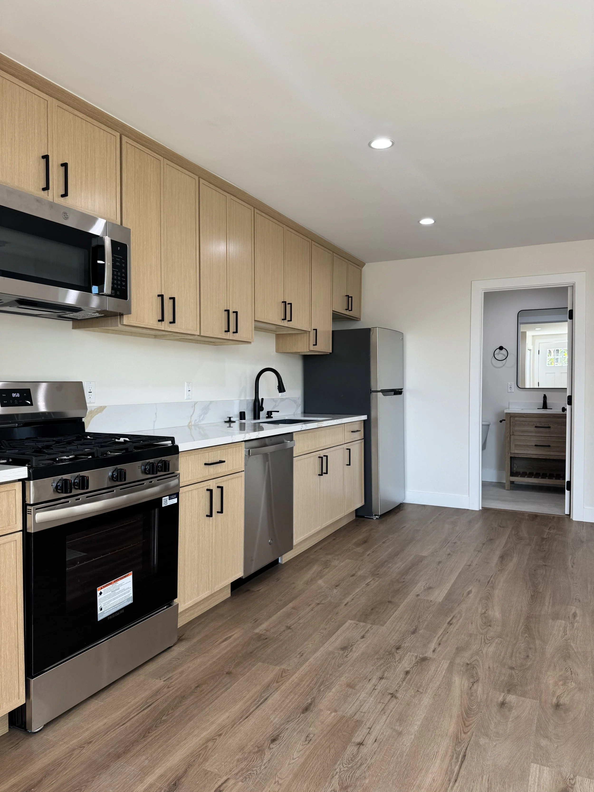 Modern kitchen with light wood cabinets, stainless steel appliances including oven, microwave, dishwasher, and refrigerator, marble backsplash, black kitchen faucet, and a doorway leading to a small bathroom with a wooden vanity and mirror.