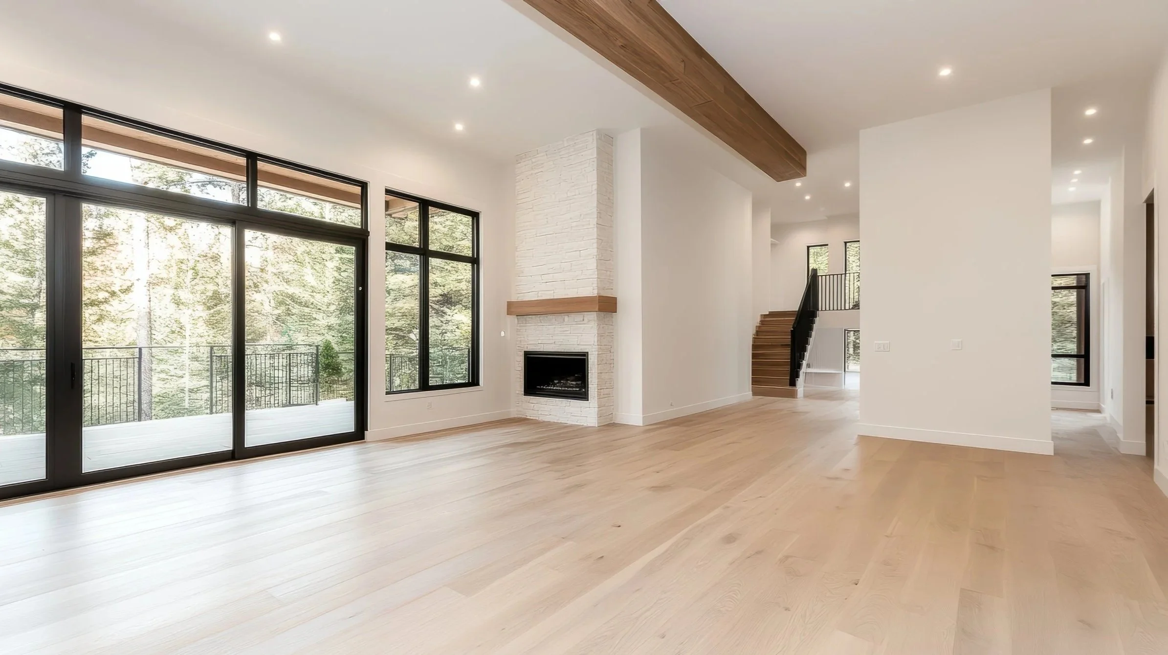 Empty living room in modern home with large windows, fireplace, wooden beam ceiling, light hardwood floors, and a staircase leading upstairs.