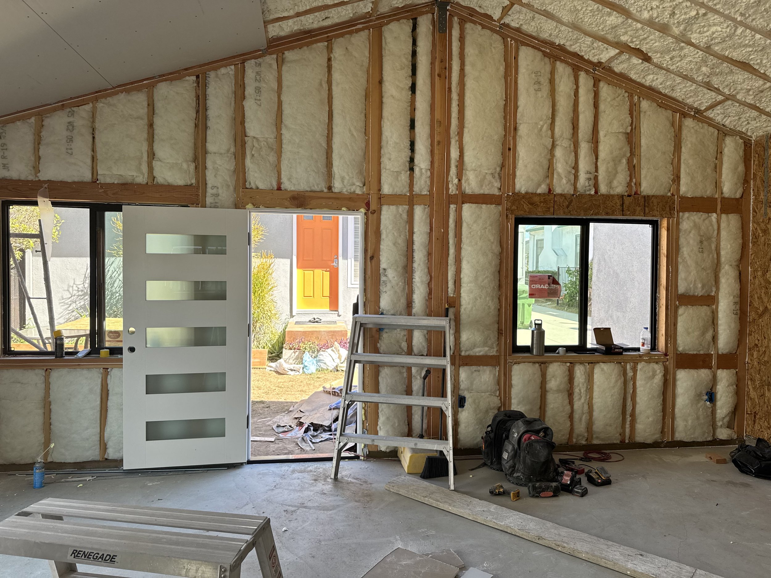 Interior view of a house under construction, showing insulation in the walls, a door opening to an outside area with a colorful door, and two windows. Construction tools and materials are on the floor.