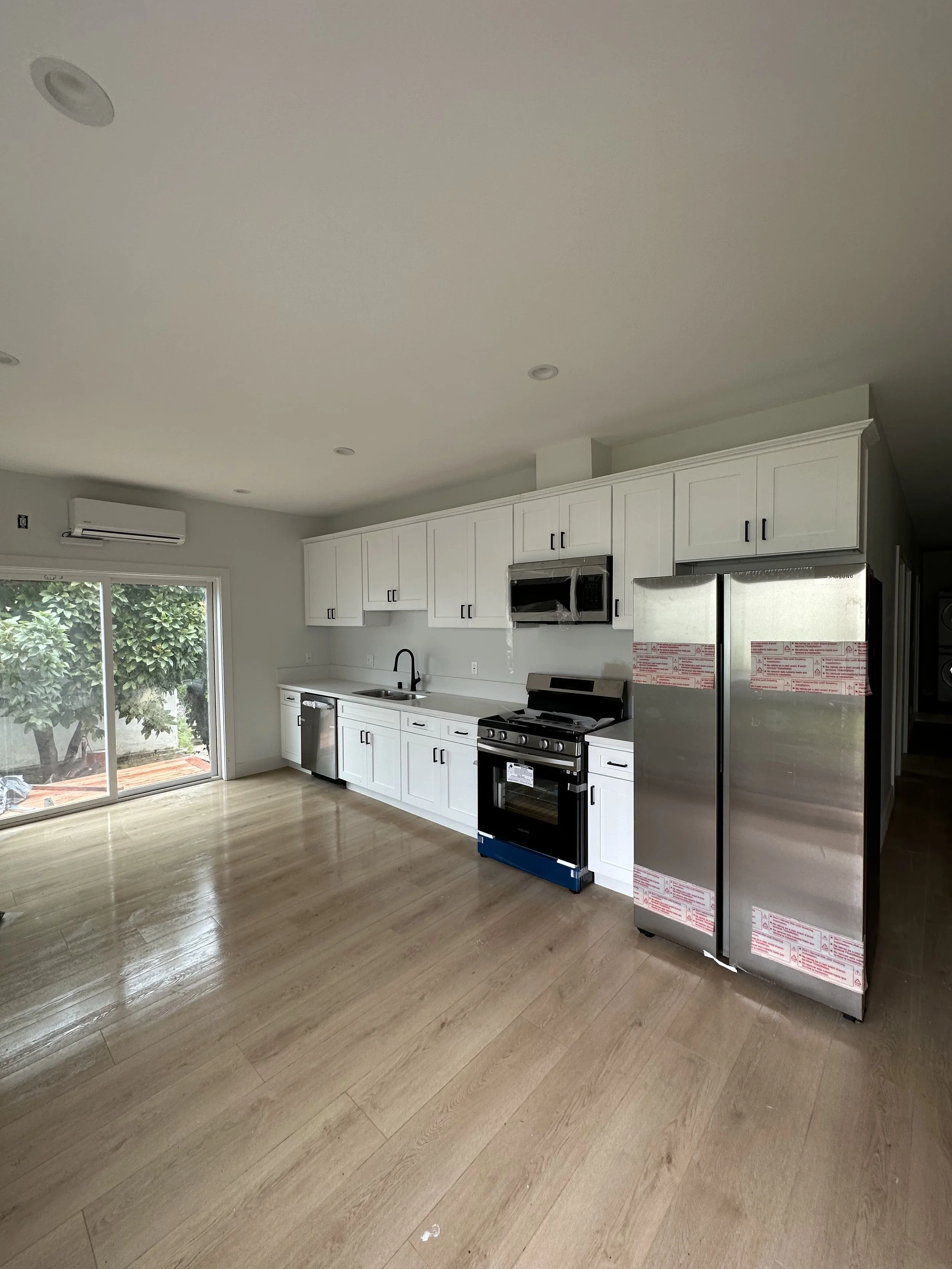A modern kitchen with white cabinets, a stainless steel refrigerator, a gas oven, and a black microwave. There is a sliding glass door leading outside, and a window unit air conditioner on the wall. The floor is light wood.